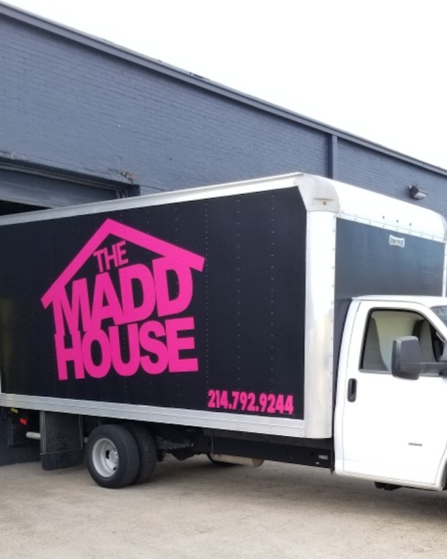 A moving truck with a black side panel displaying the logo for THE MADD HOUSE in pink, along with a phone number 214.792.9244.