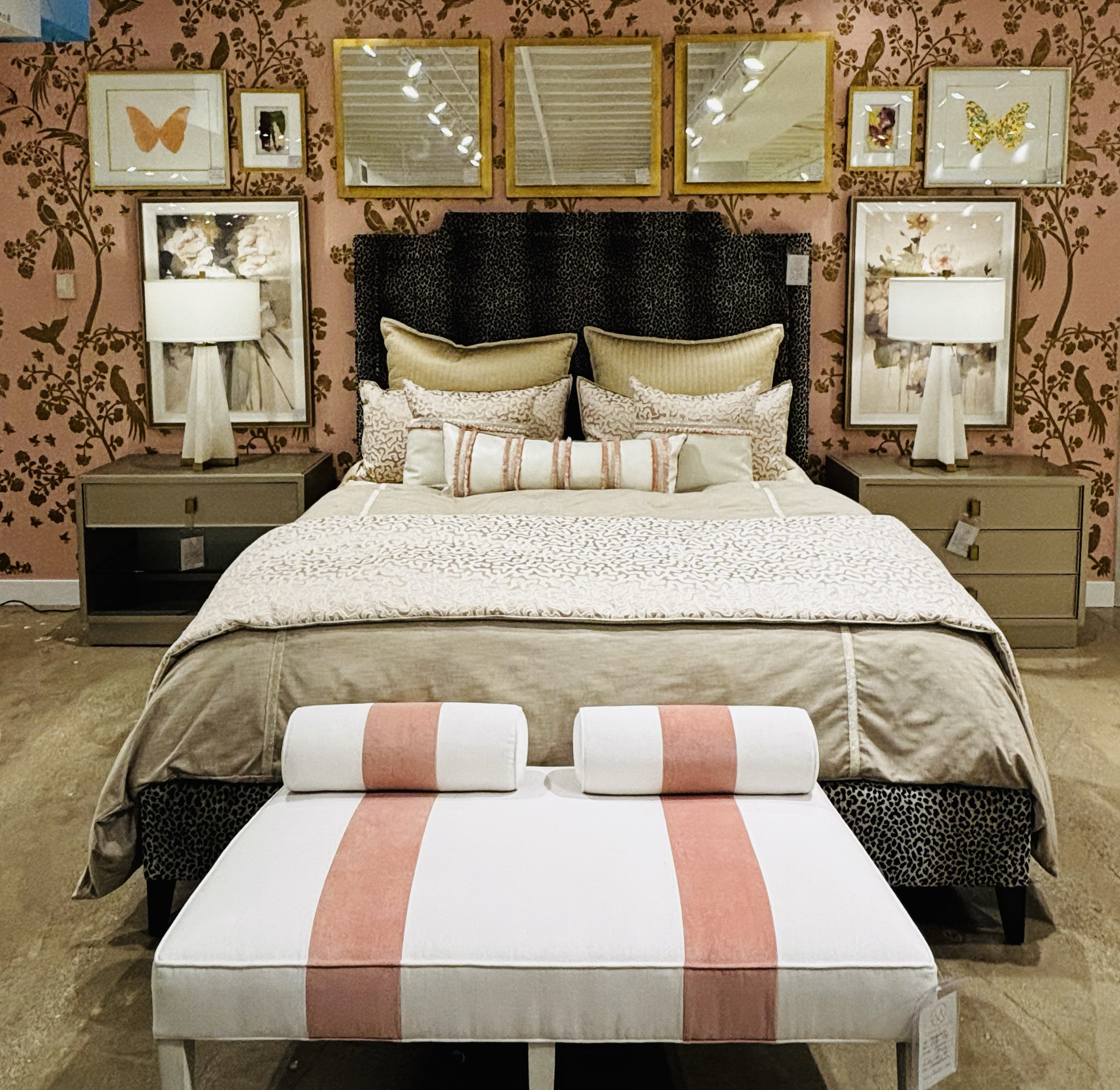 A bedroom setup featuring a bed with beige and white pillows, a black and beige headboard, and a patterned bench at the foot of the bed. The wall behind the bed is decorated with pink floral wallpaper and framed butterfly and floral artwork. There ar