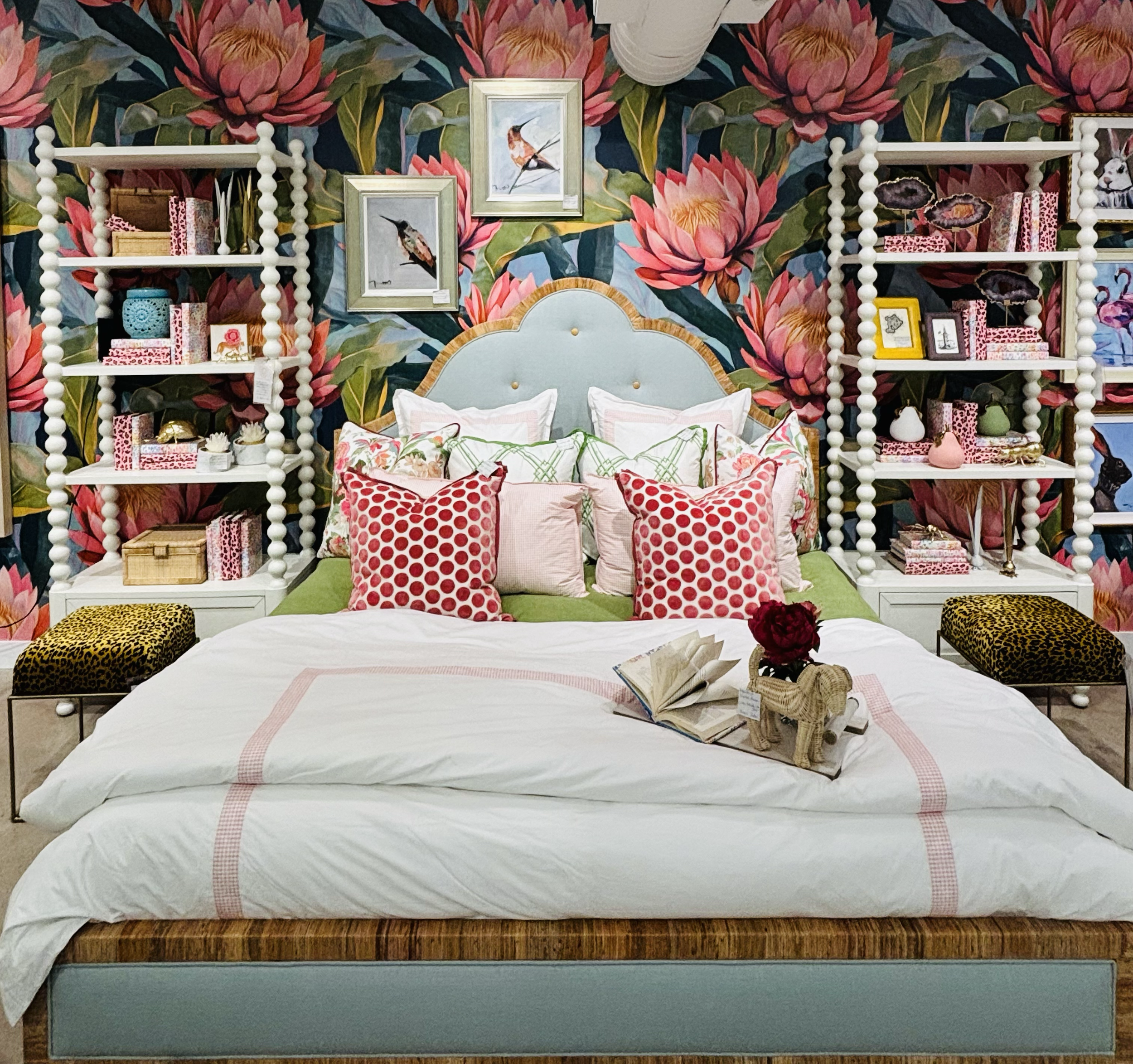 A bedroom with a bed featuring a green headboard and surrounded by colorful pillows with patterns. Behind the bed is a wall covered in floral and bird-themed wallpaper with large pink and red flowers and framed bird artwork. There are white shelves on either side of the headboard holding decorative items, books, and picture frames. A book and a woven basket with a red flower are placed on the foot of the bed.