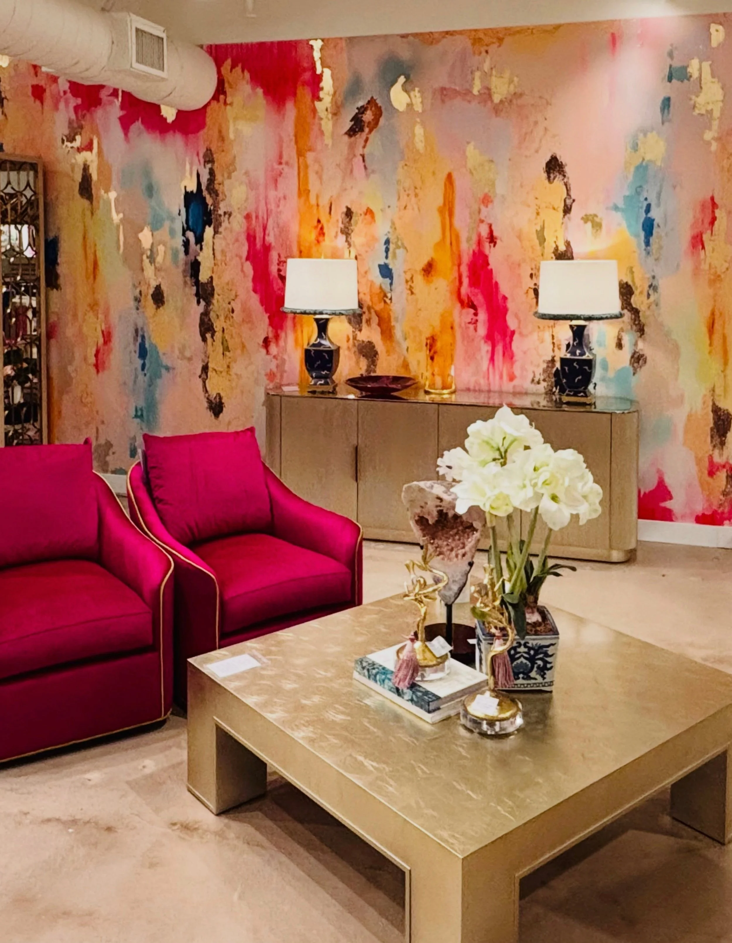 Interior of a room featuring a colorful abstract wall painting, a red velvet sofa with matching cushions, a beige coffee table with decorative items including a vase with white flowers, ornate sculptures, and a decorative box, with two black and white table lamps placed on a sideboard against the wall.