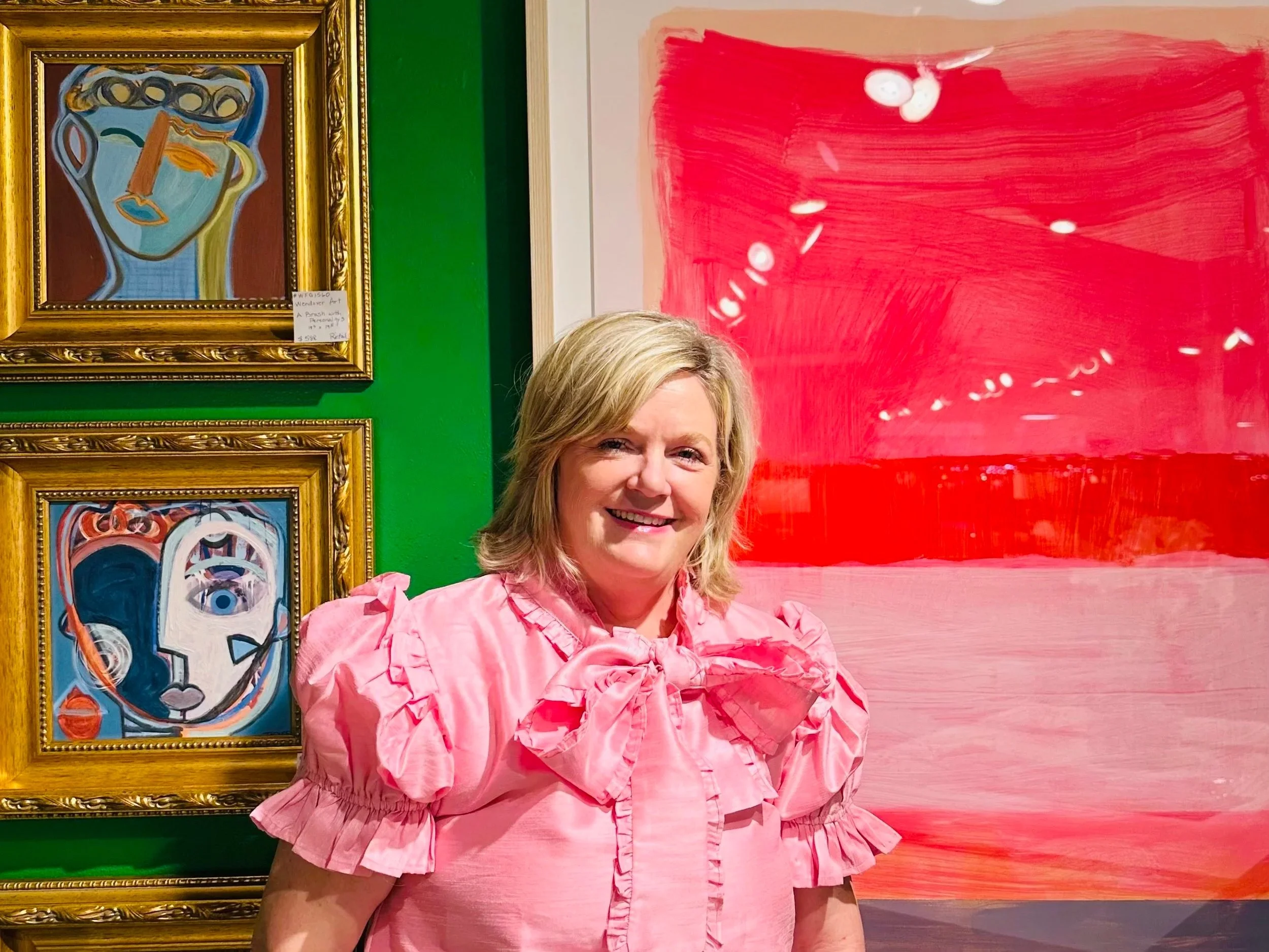 A woman with blond hair smiling, wearing a pink ruffled top, standing in front of colorful abstract artwork on a gallery wall.