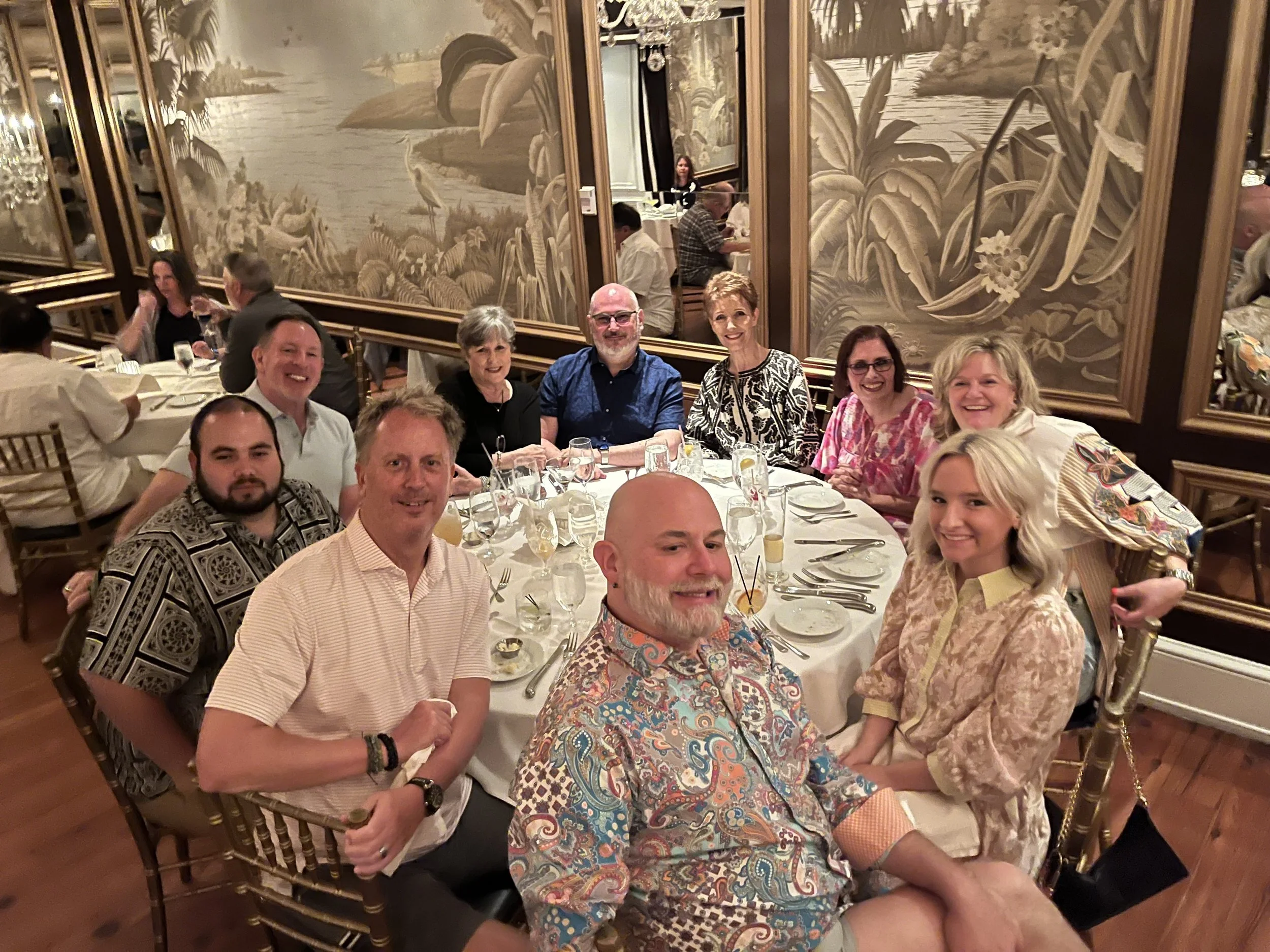 A group of nine people are gathered around a table at a formal dinner event in a decorated restaurant. The background features ornate wall art depicting a natural scene with plants and water.
