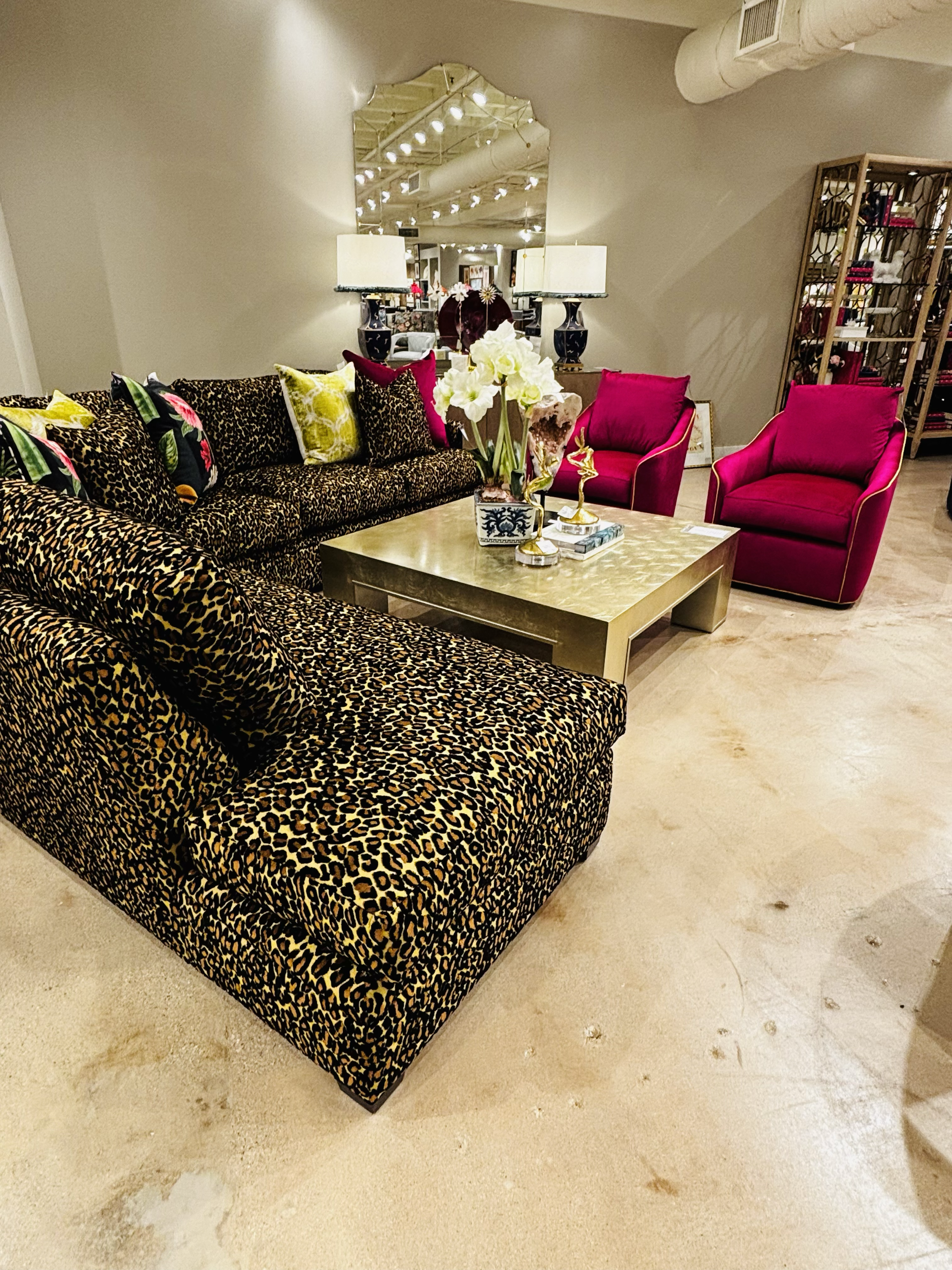 A living room with a sofa covered in leopard print fabric, two pink armchairs, a central coffee table with a floral arrangement, and a mirrored wall behind the sofa with lamps on either side. There is a shelving unit on the right side and a textured beige floor.