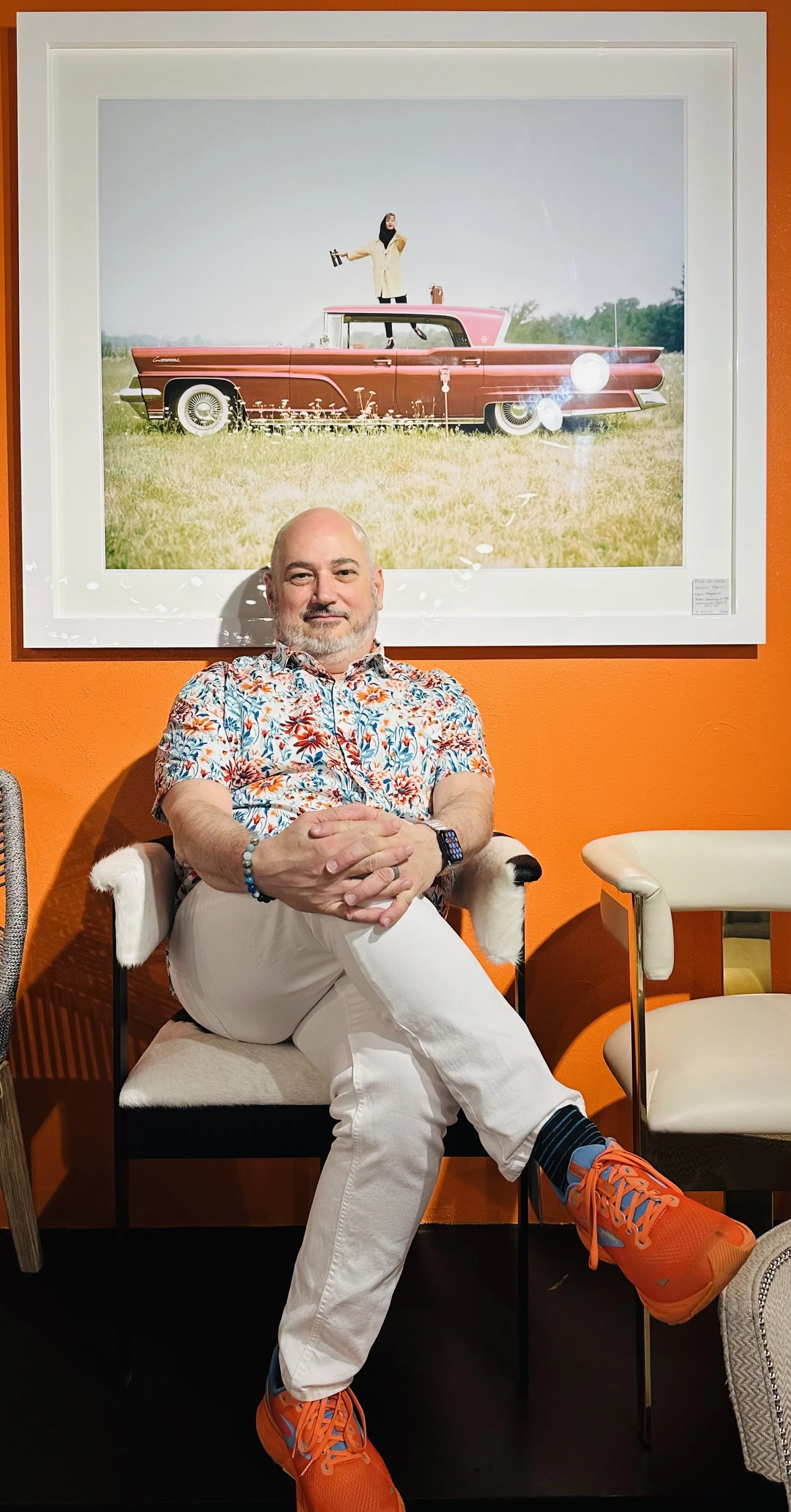 A man sitting with legs crossed, wearing orange sneakers, a floral shirt, white pants, and a smartwatch, smiling in front of a framed photo of a vintage car with a woman standing on top, holding items, in a grassy field.