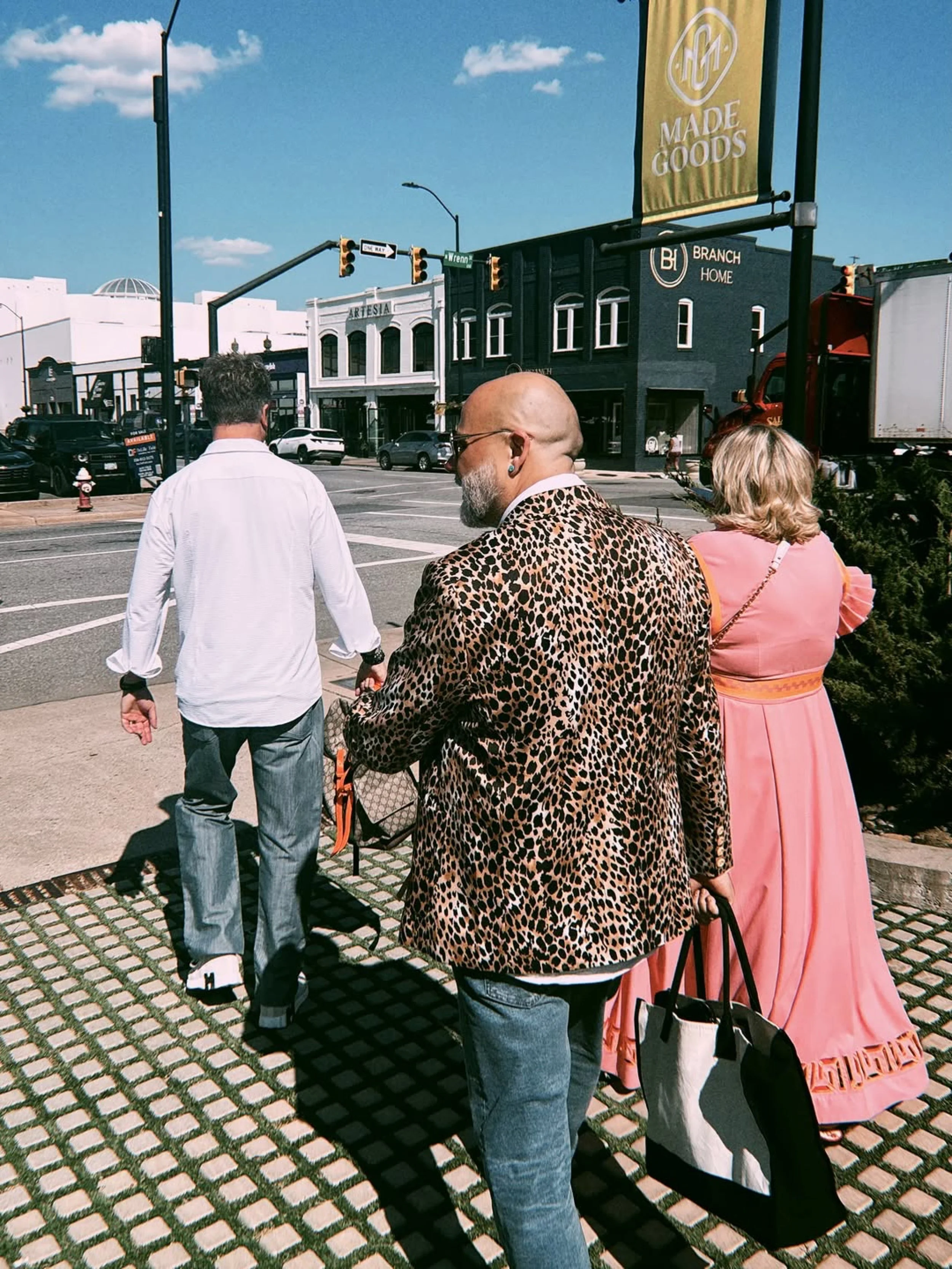 Three people are standing on a city sidewalk at a crosswalk, with storefronts and cars visible in the background. It is a sunny day with a blue sky.