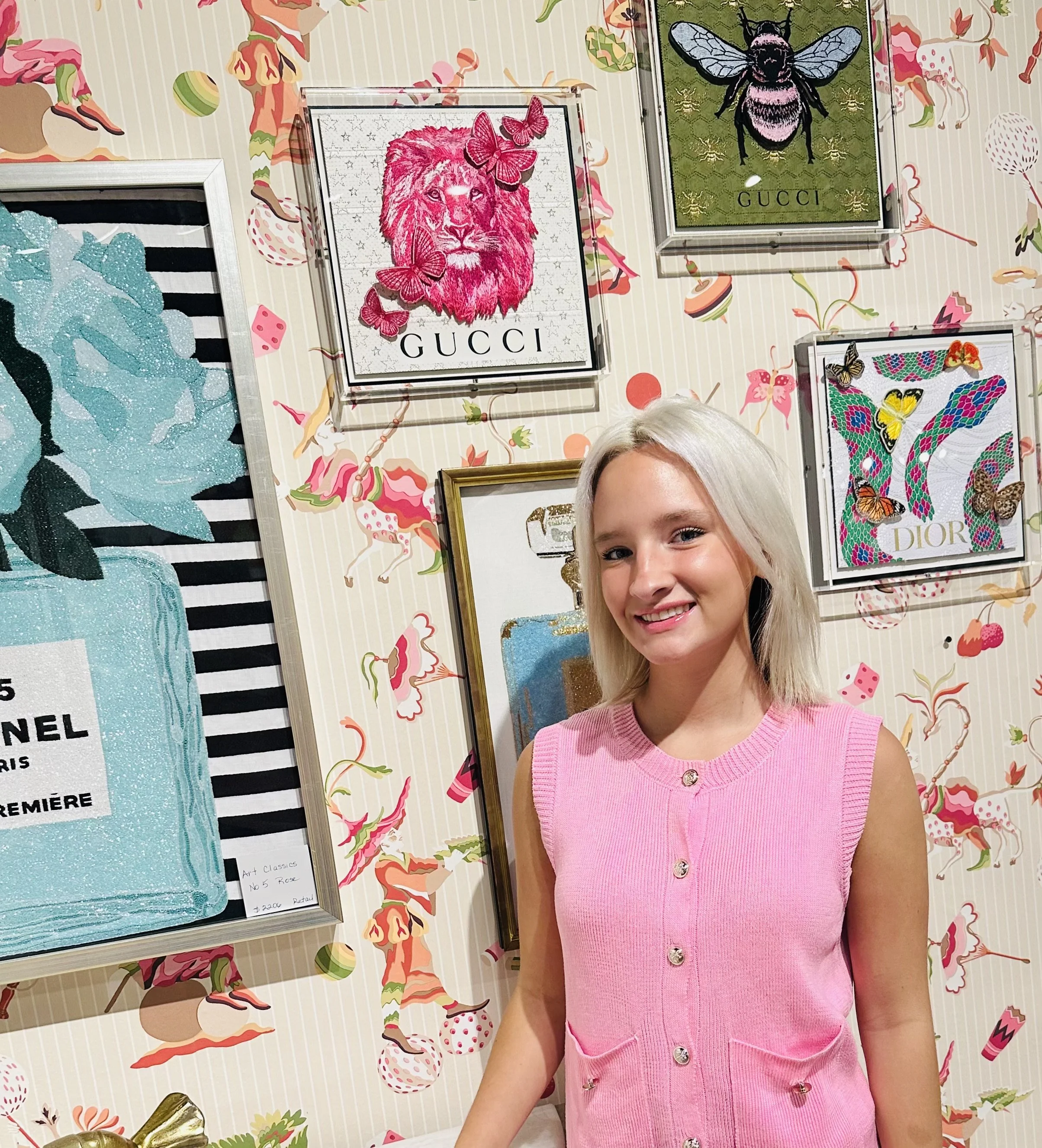 A woman with blonde hair wearing a pink sleeveless top, smiling, standing in front of colorful framed artwork on a wall with a whimsical animal and floral pattern.