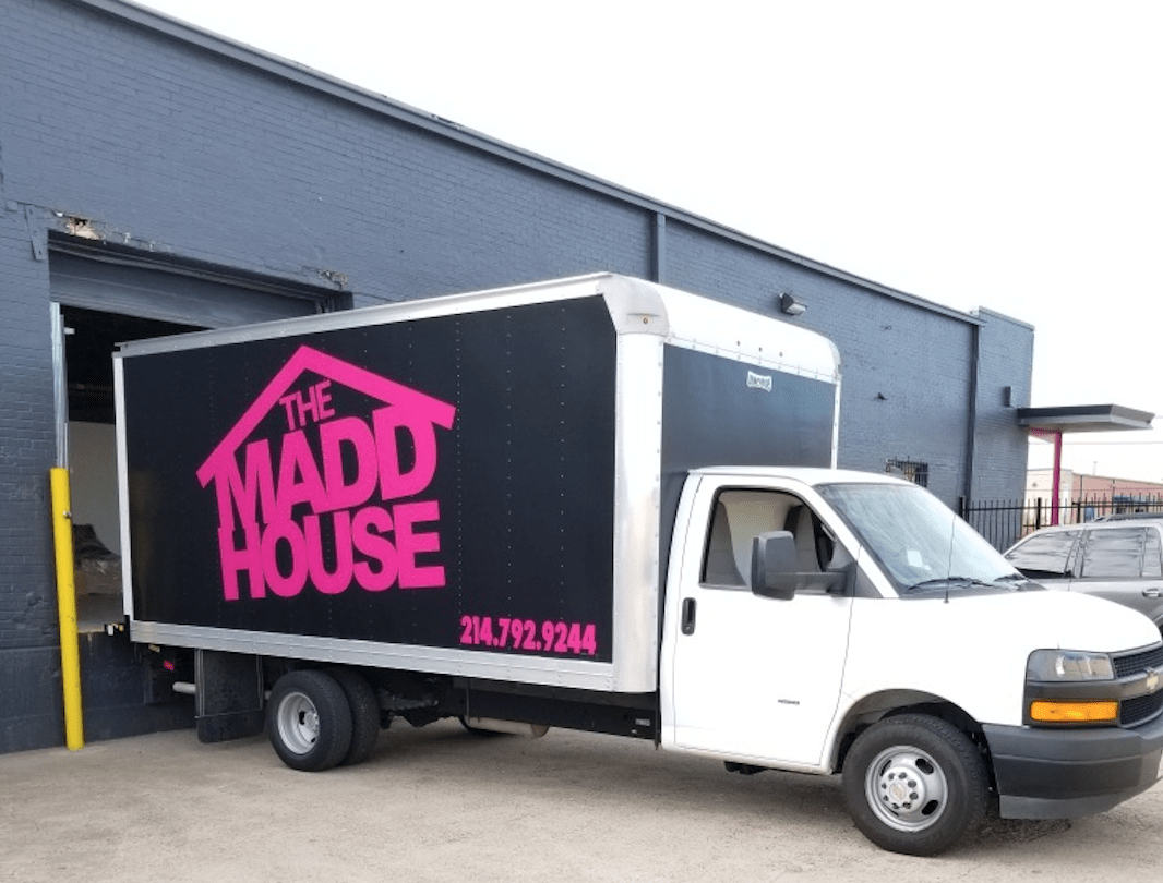 White moving truck with black and pink logo that reads 'The Madd House' and a phone number, parked outside a dark gray building.