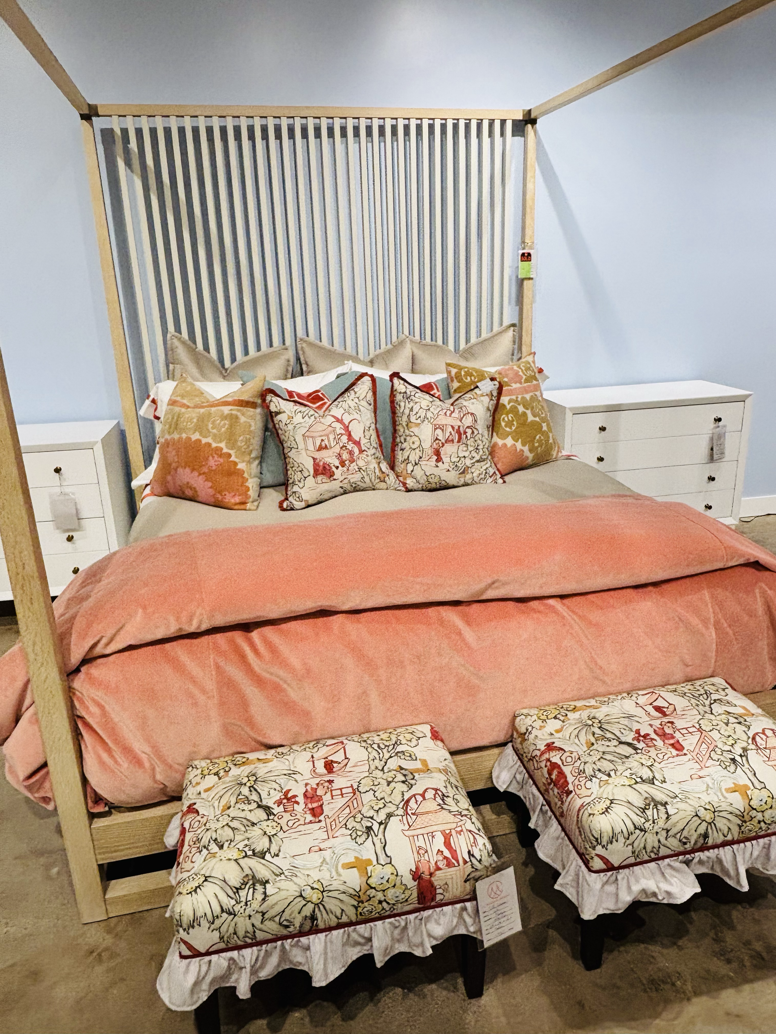 A four-poster bed with a carved wooden frame, beige and floral patterned pillows, peach-colored bedding, and a white skirt, flanked by two white nightstands with drawers, in a room with light blue walls and a beige carpet.