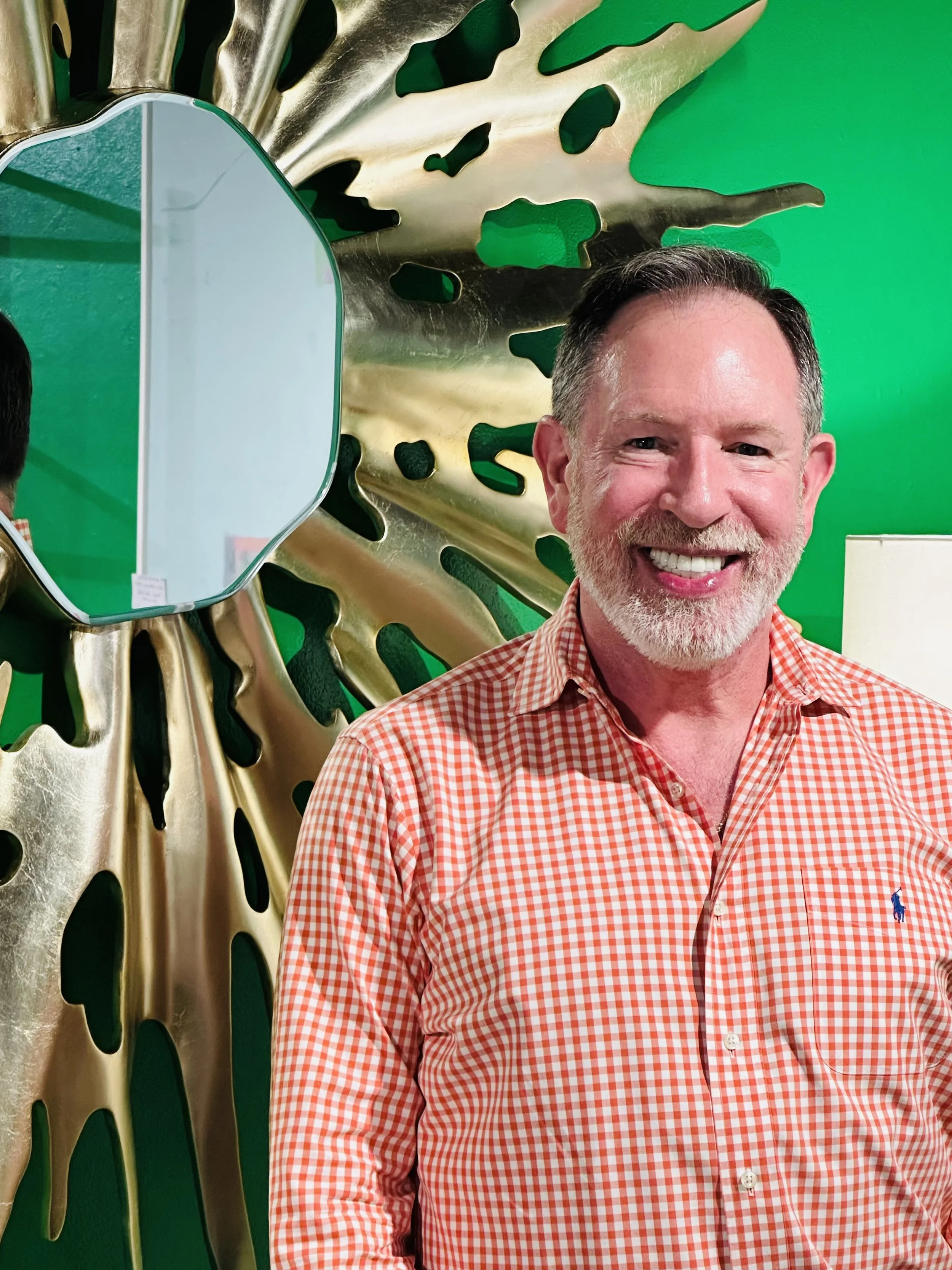 A man with a beard and short hair wearing an orange and white checkered shirt, smiling in front of a decorative wall with metallic and green designs.