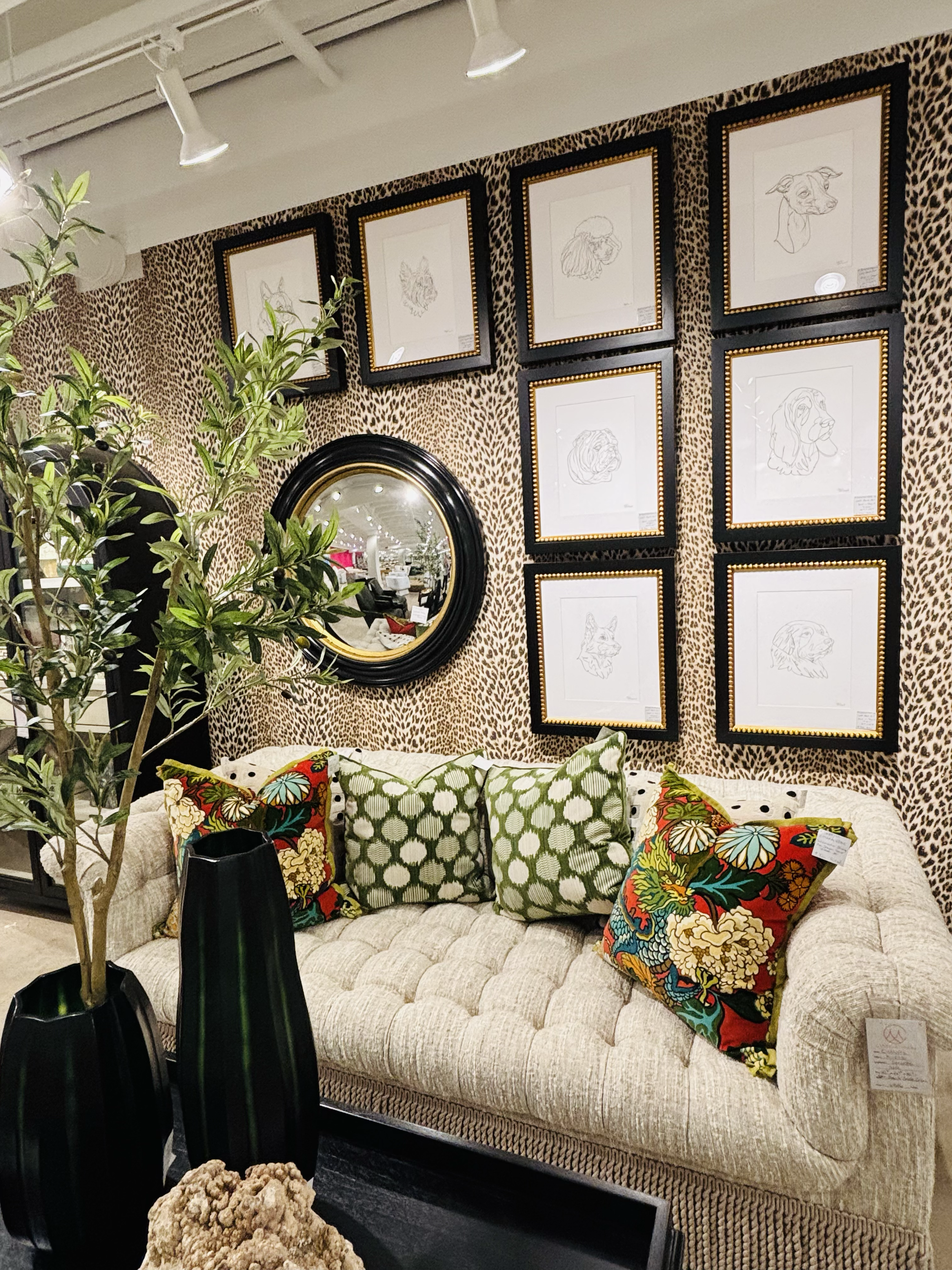 A beige tufted sofa with colorful patterned cushions in front of a leopard print wall. Above, a series of framed dog and wolf line art drawings hang on the wall. There are two tall black vases with green plants nearby, and a large mirror reflects par