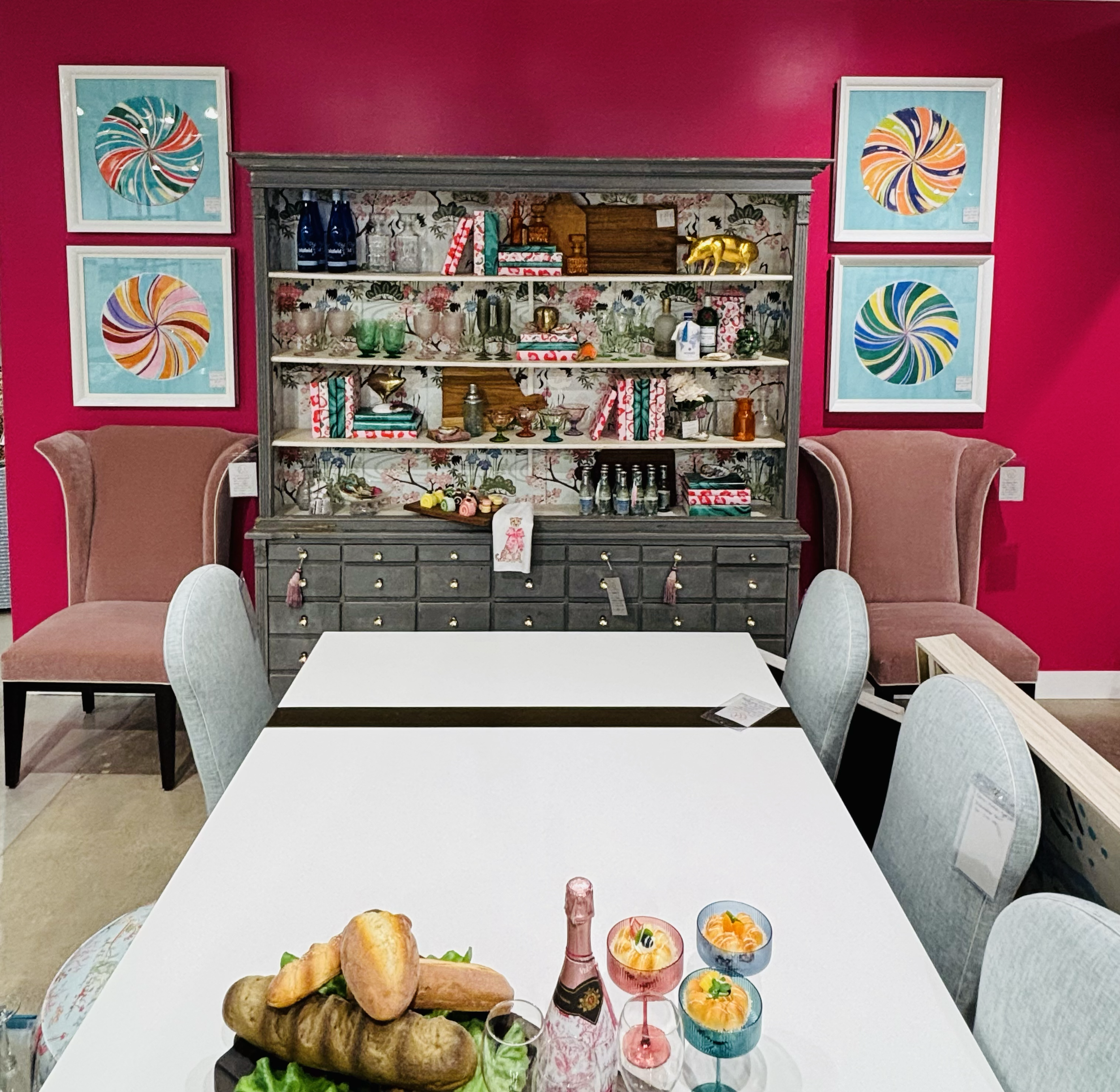 Table with food and drinks, including bread, pastries, colorful cocktails, and a pink bottle, in a bright dining setting with pink walls, floral wallpaper, and framed colorful spiral art.
