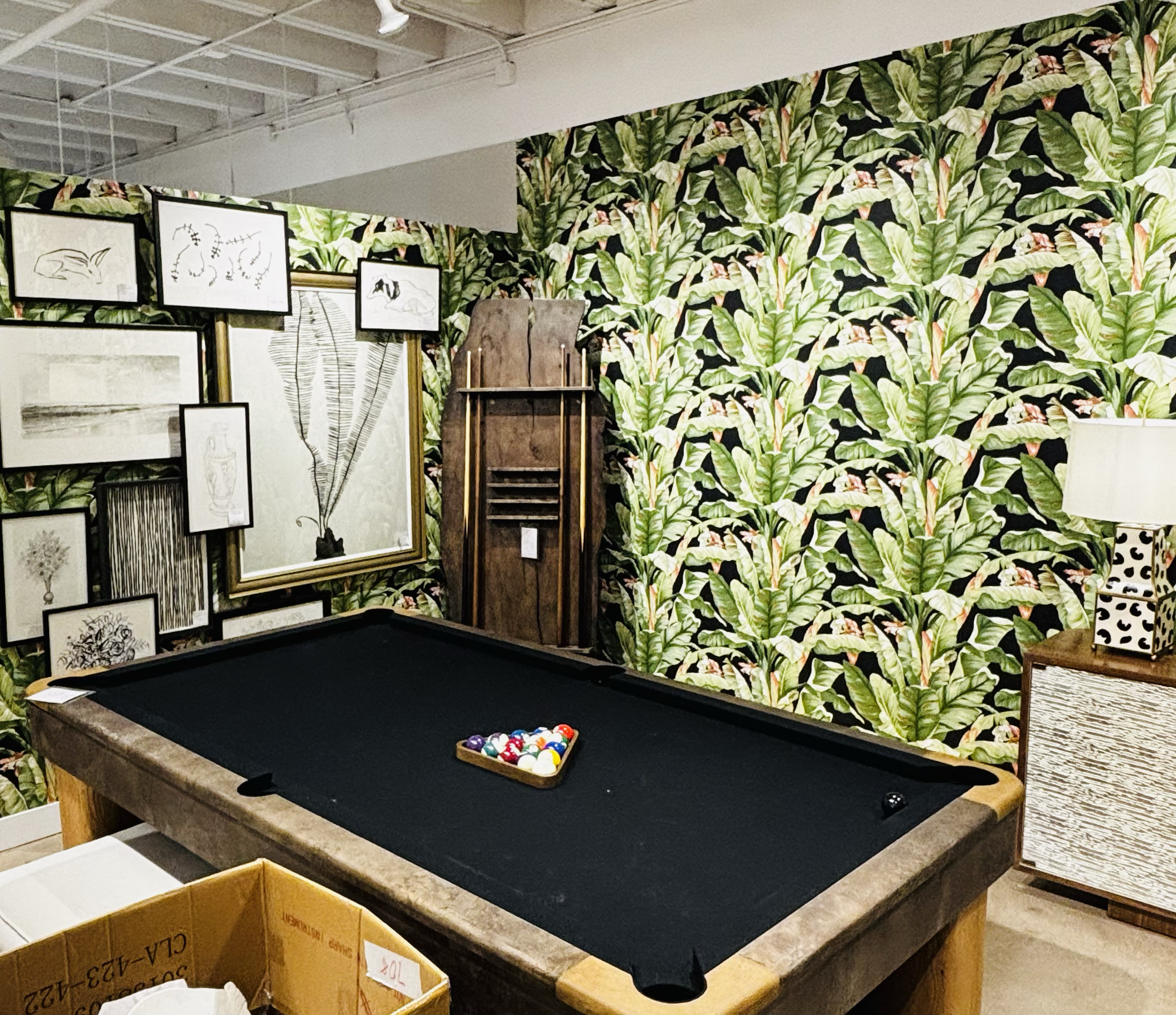 Indoor room with a pool table, framed artwork on a rack, and a vibrant tropical leaf patterned wallpaper.