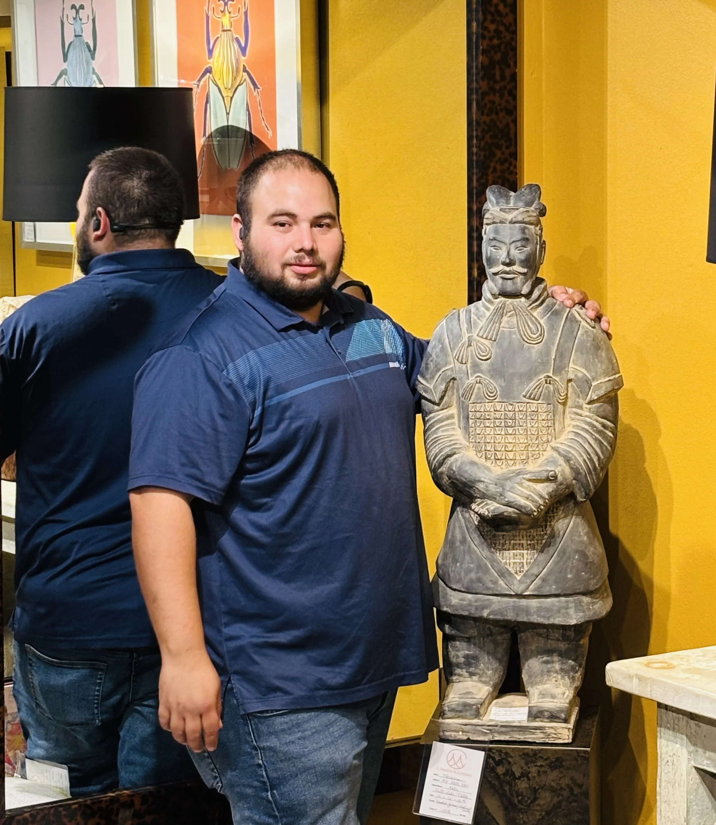 A man with a beard in a blue shirt and jeans standing next to a stone statue of an ancient warrior or king, with a yellow wall behind them.