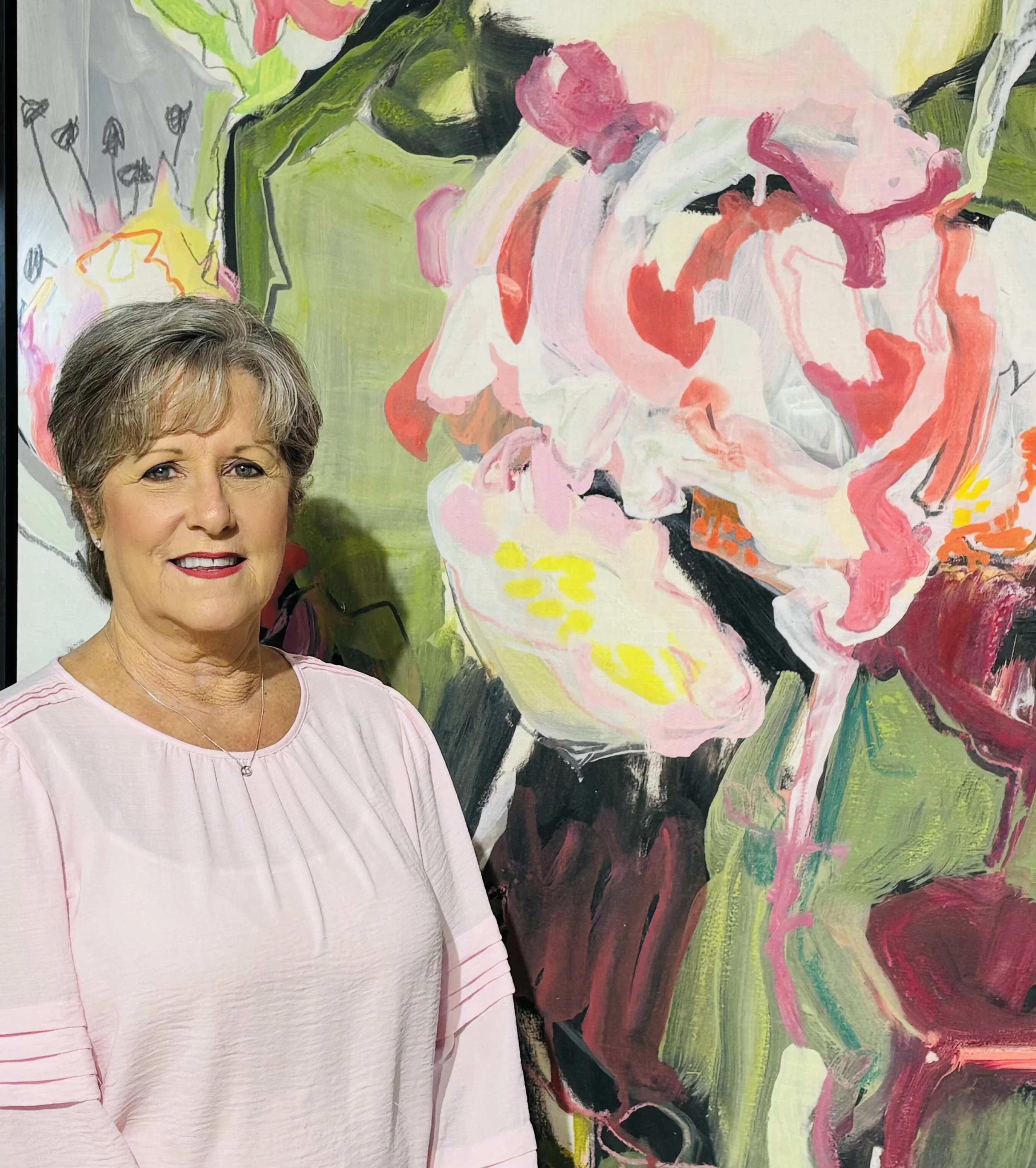 A woman with short gray hair and wearing a light pink shirt is standing in front of a colorful abstract painting with pink, white, yellow, green, and black paint strokes.