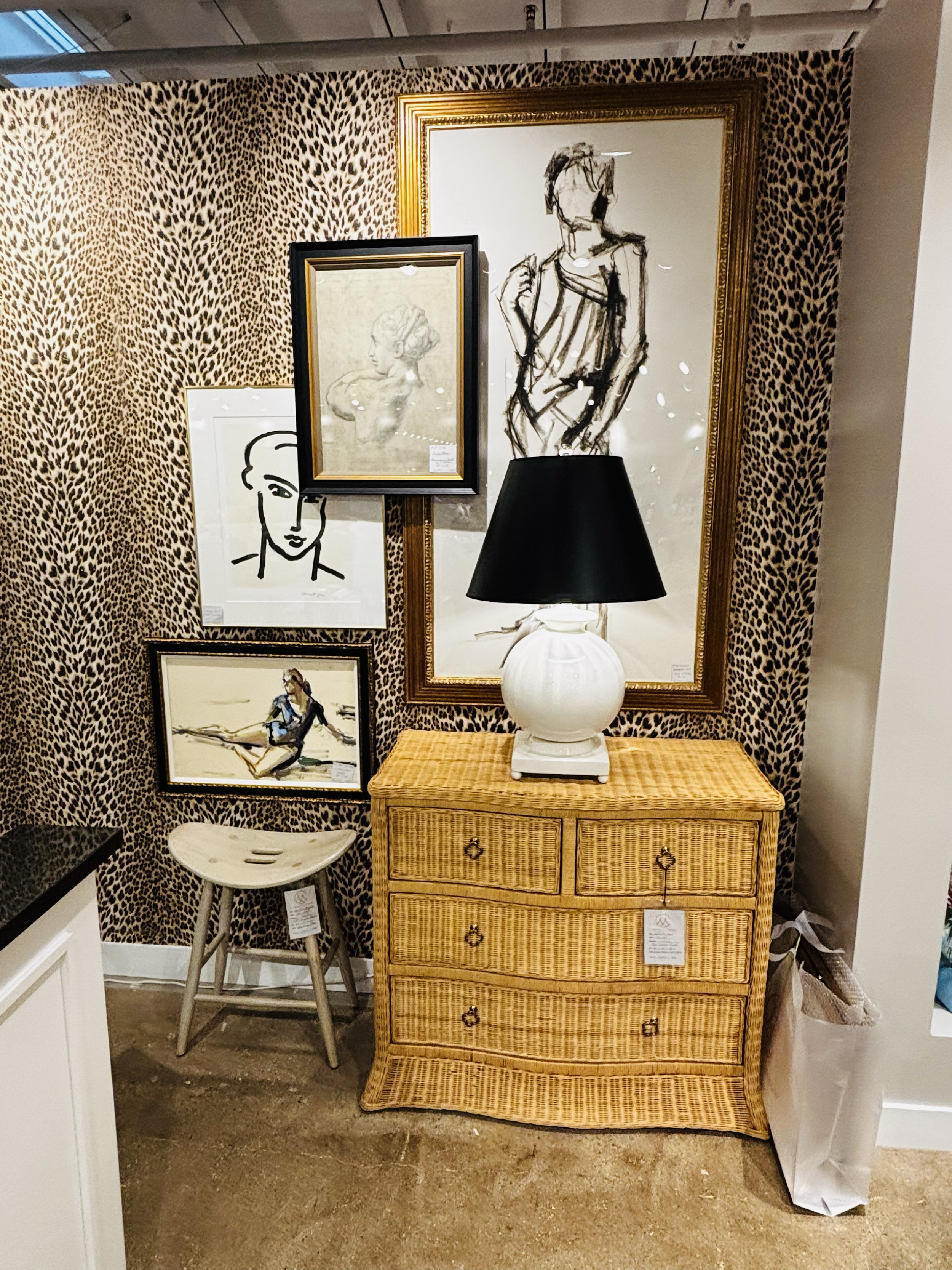 A wooden wicker dresser with a white table lamp on top, positioned in front of a large mirror with a gold frame. The wall behind features animal print wallpaper, and several framed art pieces are displayed around the mirror.