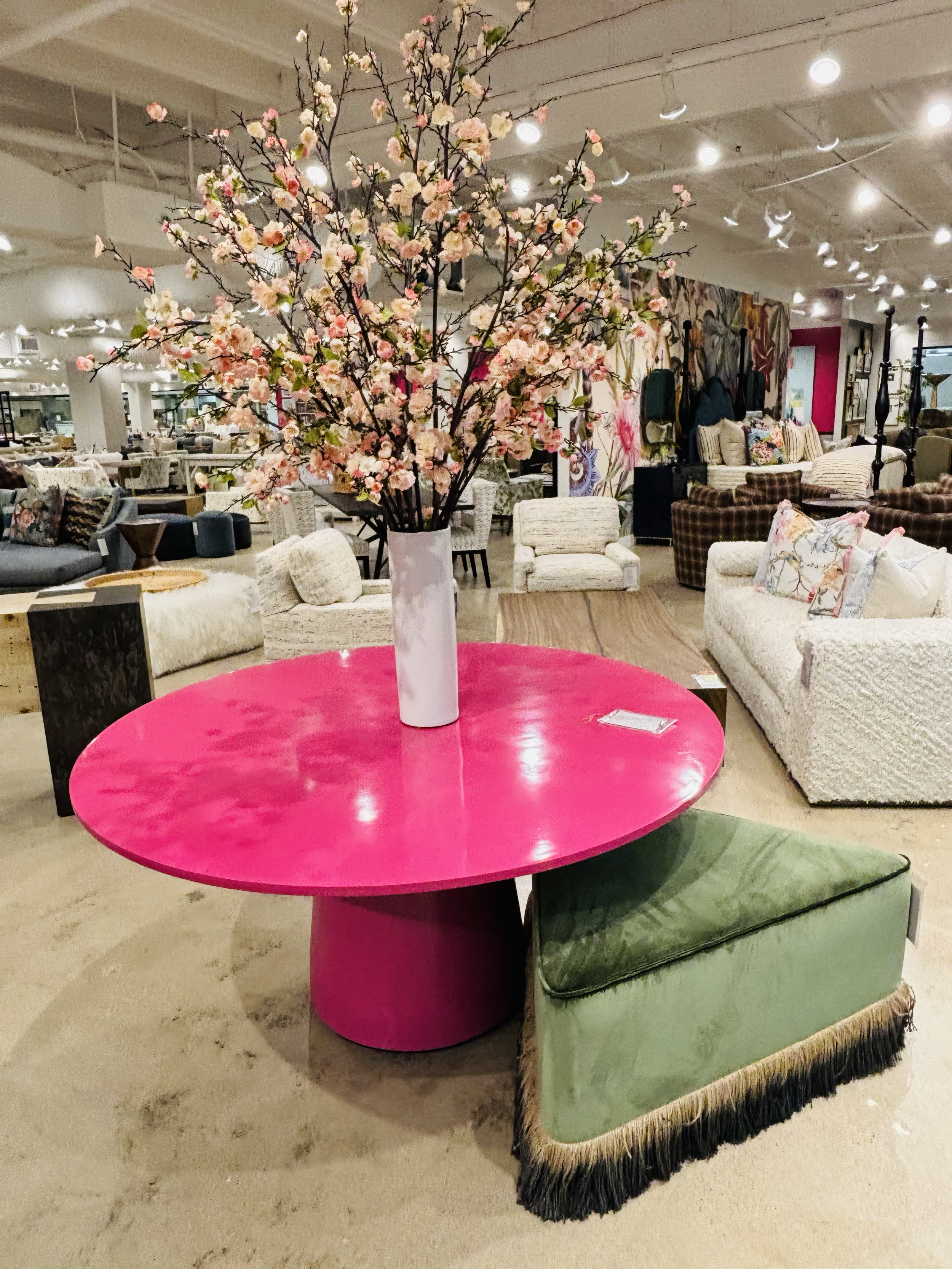 A bright pink round table with a white vase holding cherry blossom branches, surrounded by various chairs and sofas in an indoor furniture store.