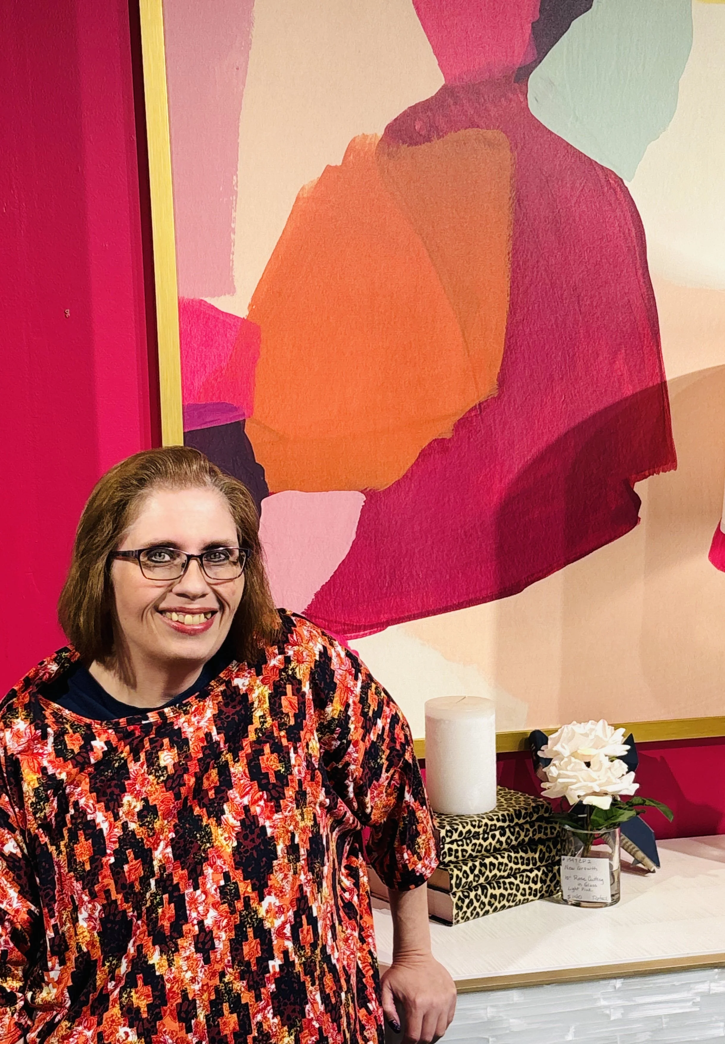 A woman with glasses and shoulder-length brown hair, wearing a colorful patterned dress, smiling at the camera, standing in front of a table with various items and a colorful abstract wall art in the background.