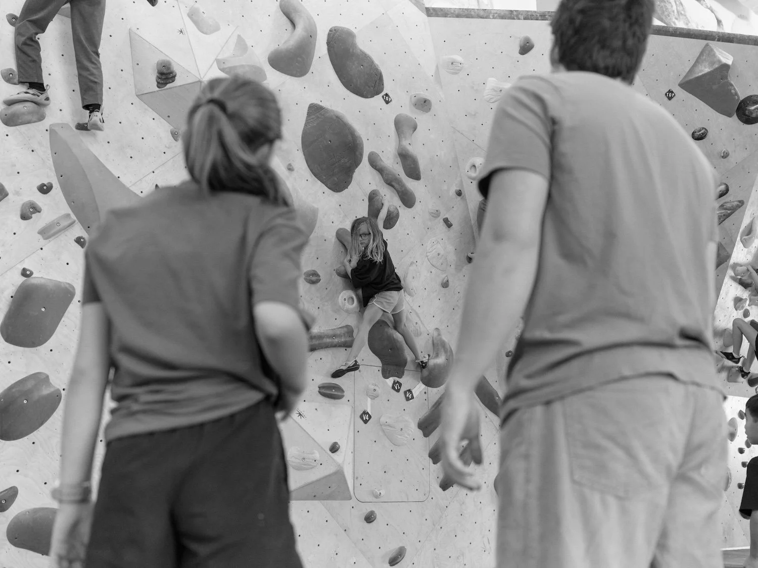 Black and white image of a young boy climbing on a rock climbing wall with two adult instructors in the foreground.