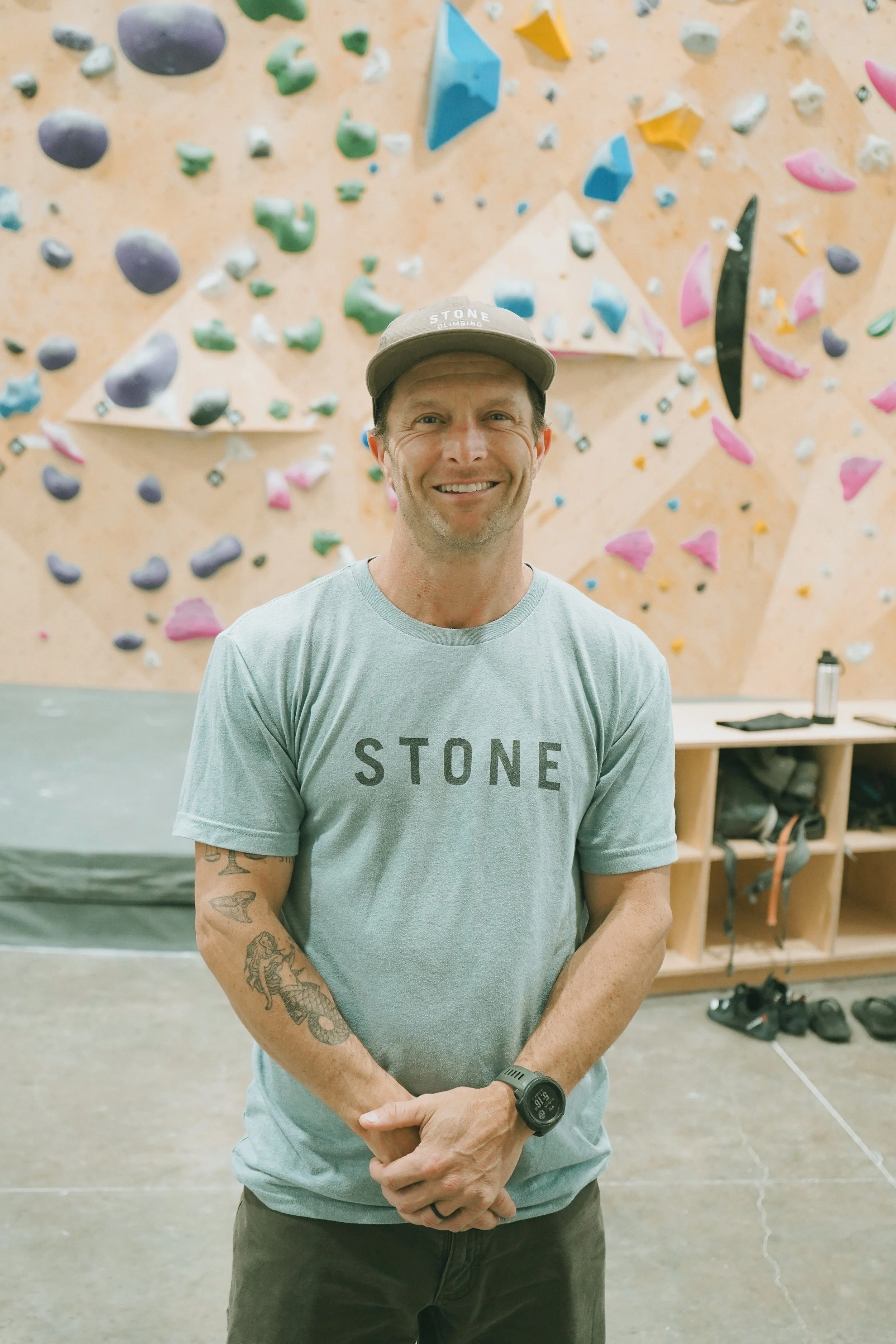 A man standing in front of an indoor climbing wall, smiling, wearing a gray T-shirt with the word 'STONE' and a hat that says 'STONE CLIMBING'.