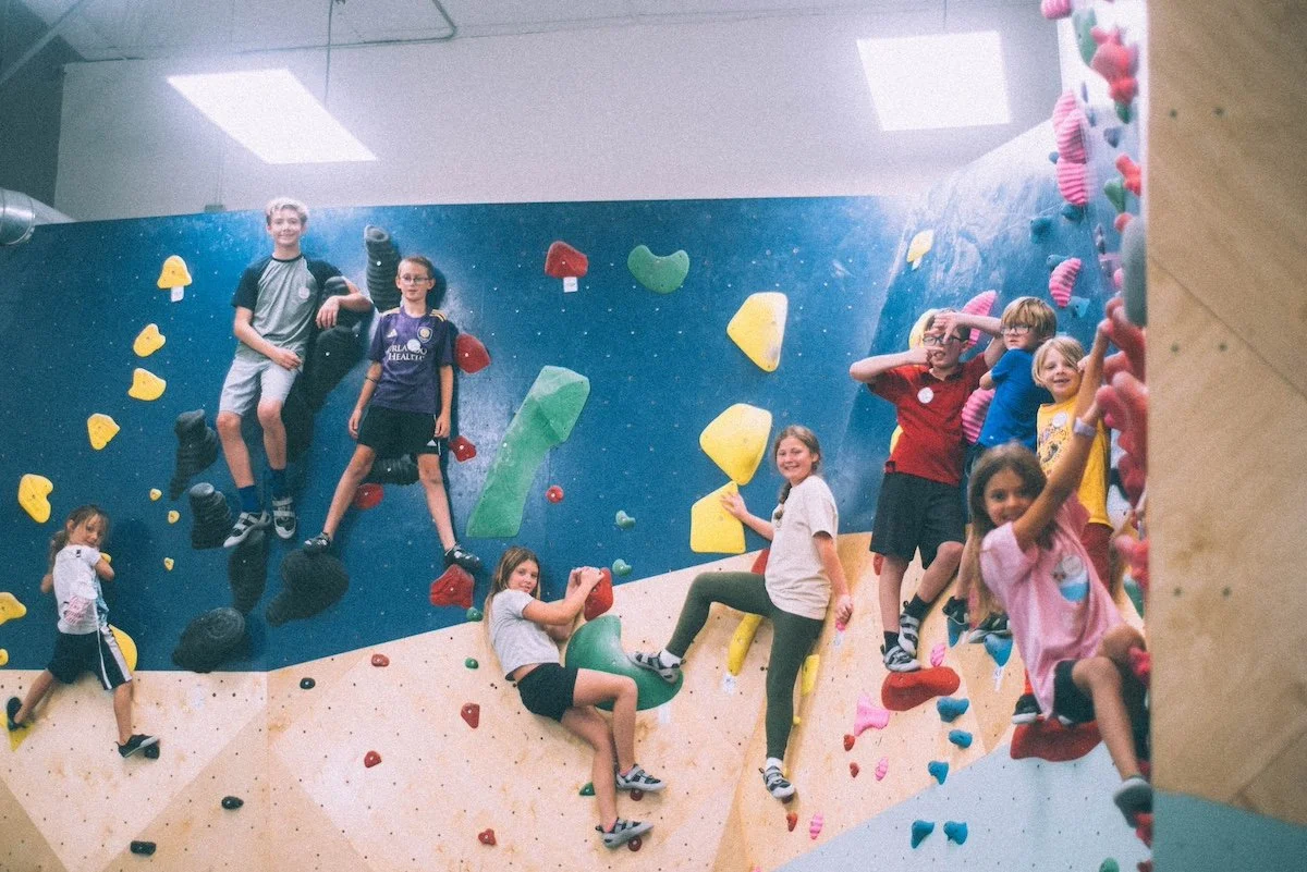 Children climbing and playing on an indoor bouldering wall with colorful holds in a gym.