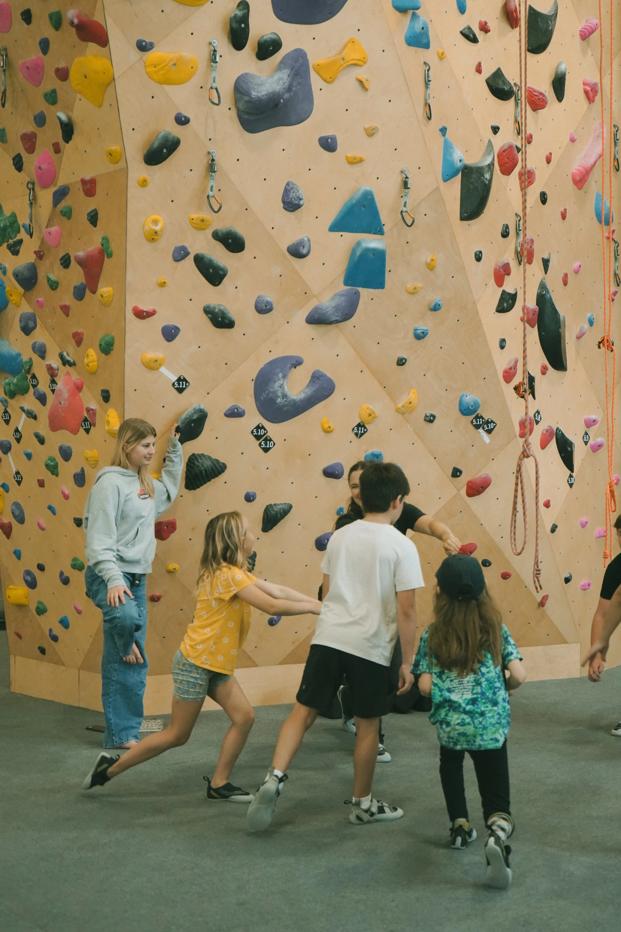 A group of children playing and running inside an indoor rock climbing gym. The climbing wall behind them is covered with colorful holds and ropes.