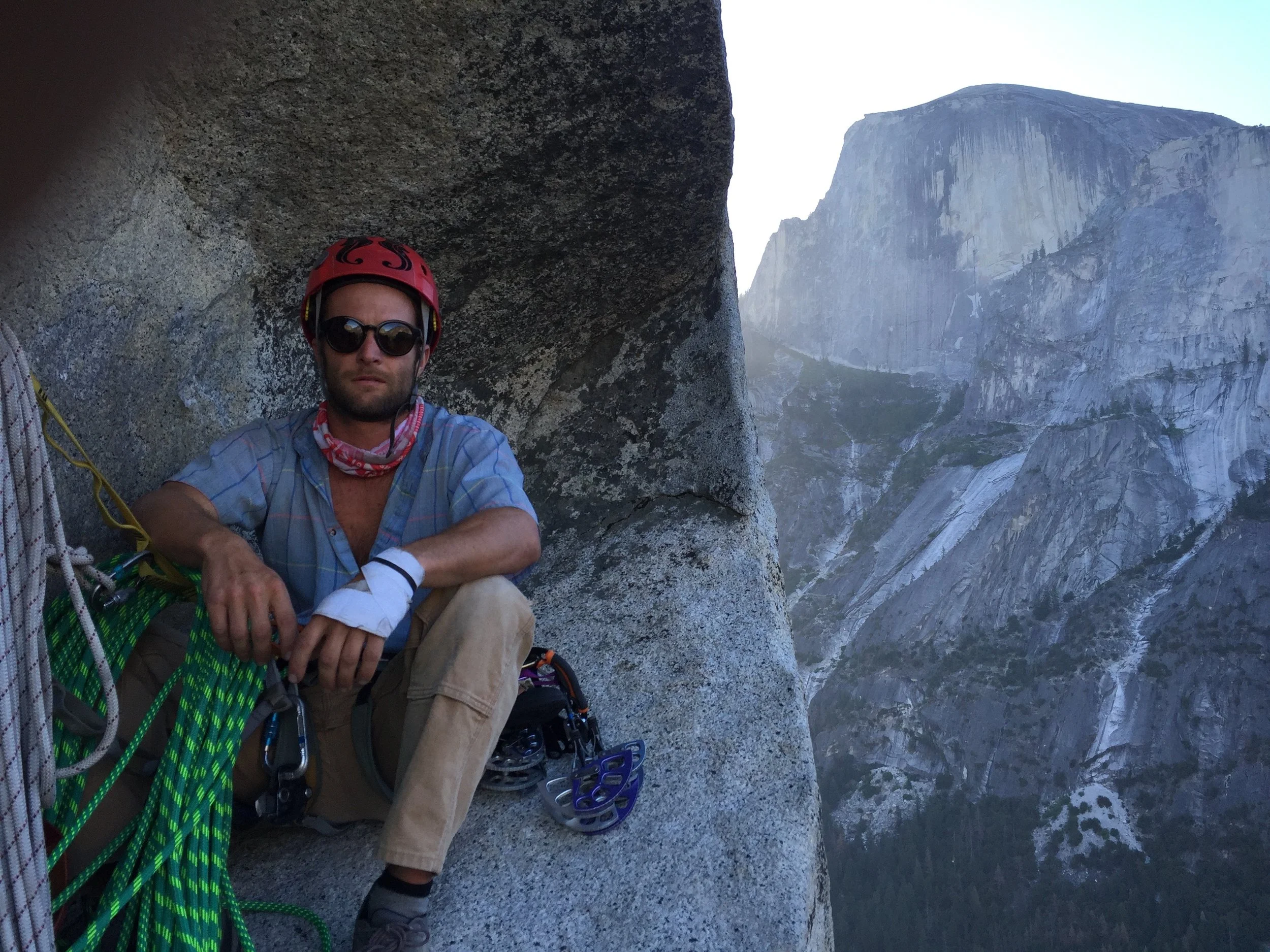 A man wearing a red climbing helmet and  blue shirt, sitting on a rock face during a rock climbing or mountaineering activity, with half dome and forest in the background.