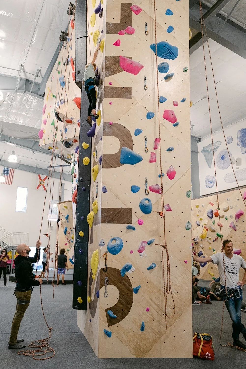Instructors belaying kids on a rope climbing wall at an indoor climbing gym with colorful holds and bouldering walls in the background.