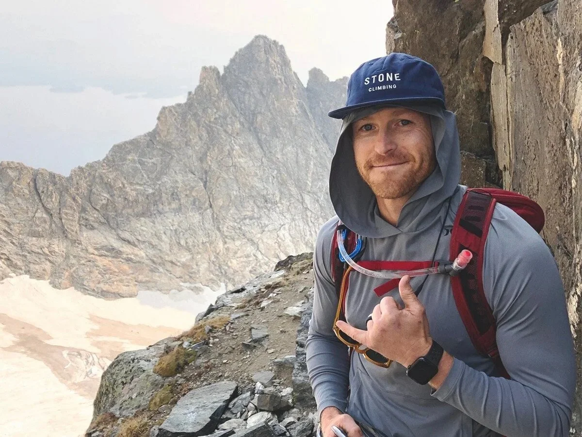 A man in outdoor gear smiling and making a rock on sign in a mountain landscape with rugged rock formations and snow patches.