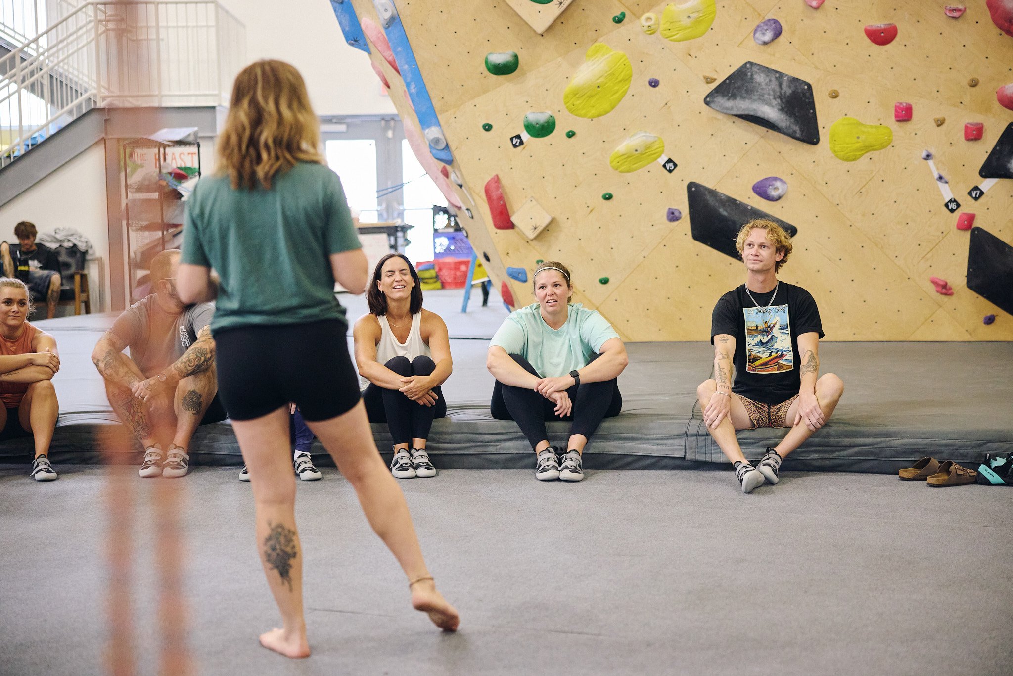 Climbing instructor talking to students taking part in a team building experience at a rock climbing gym with climbing walls in the background.