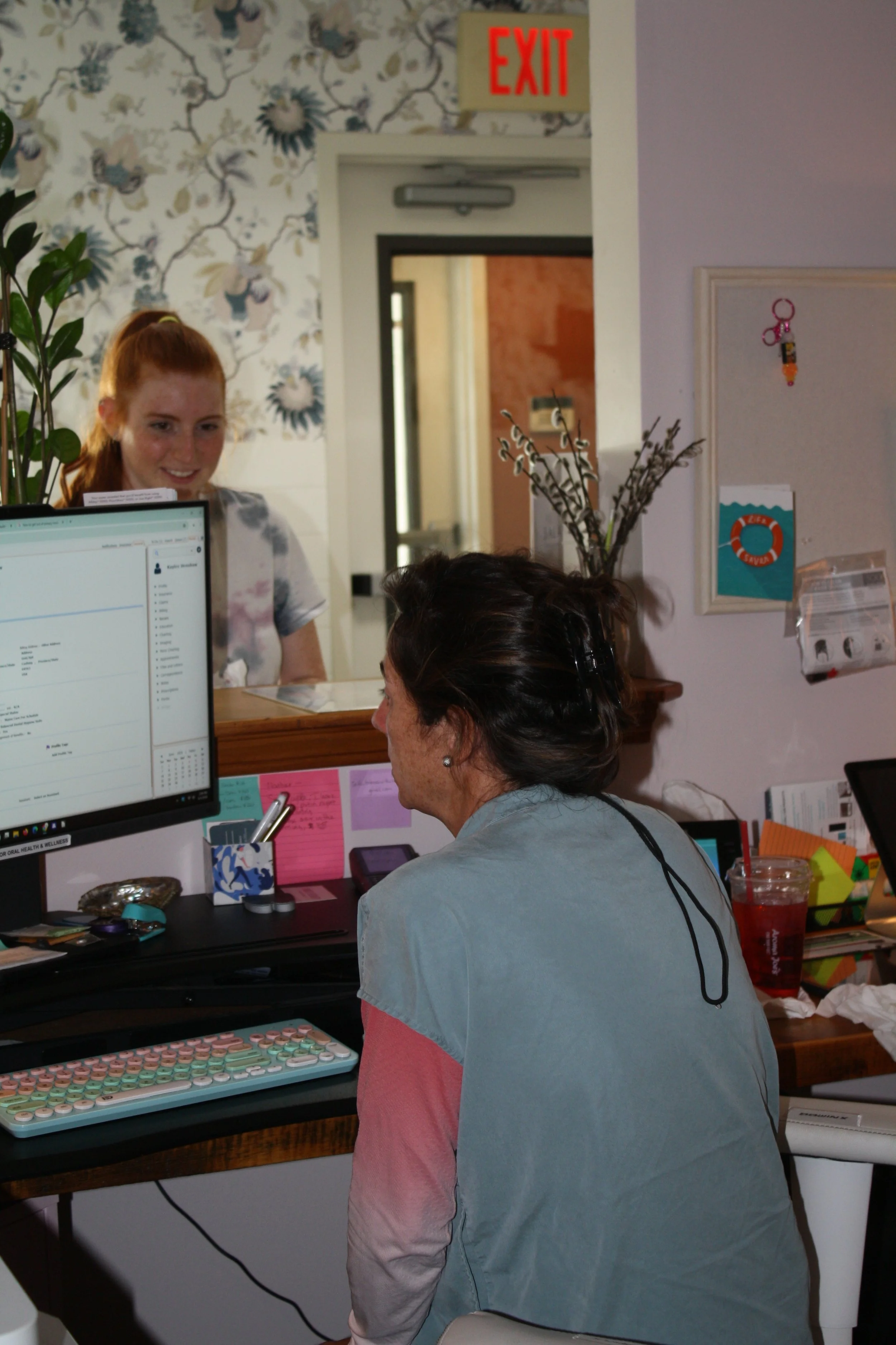 Owner and Founder, Kala French, sits at a desk in a medical or dental office, looking at a computer monitor. Behind her, a reception area is visible with a woman with red hair smiling in a windowed partition.