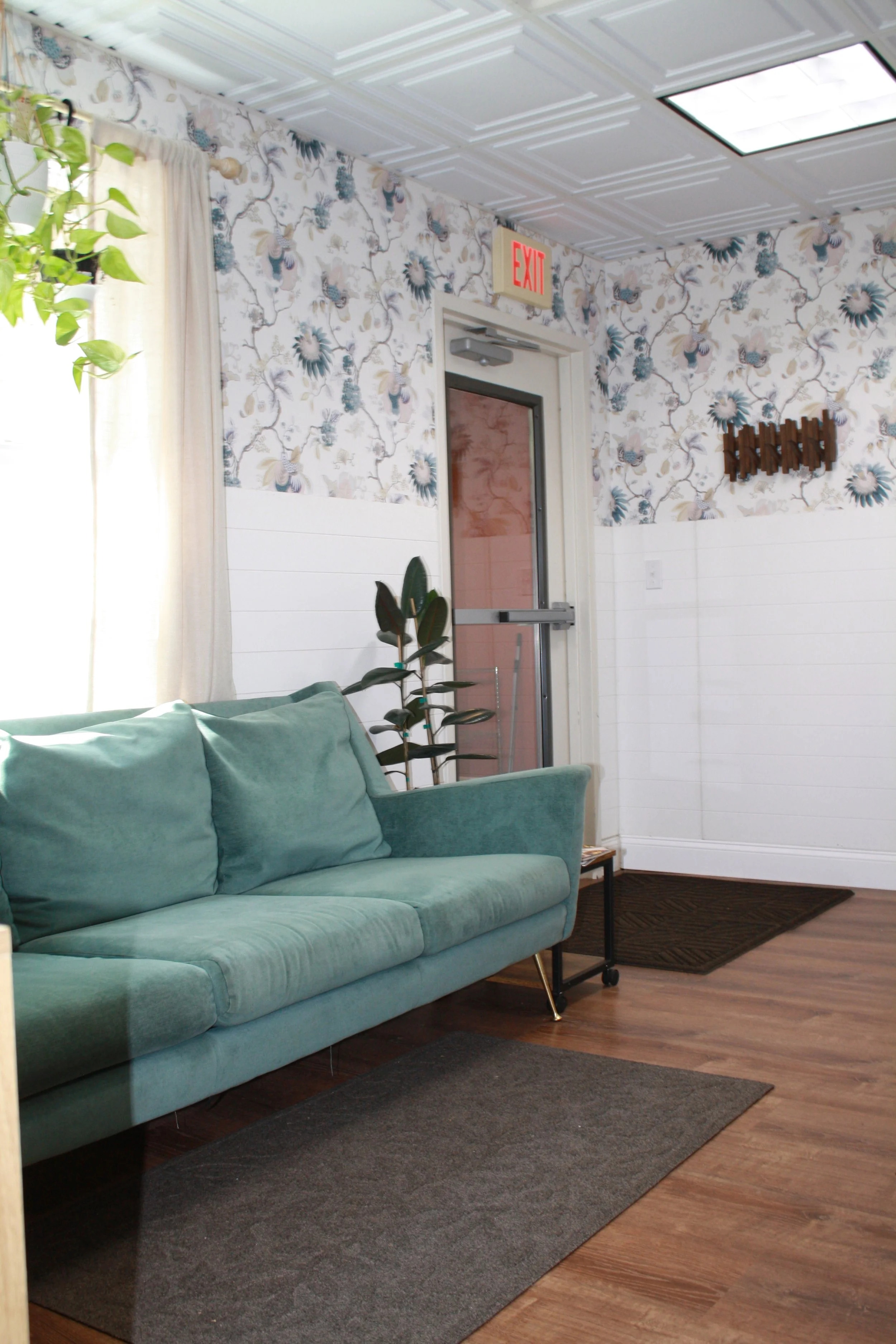 Interior of a room with a teal sofa, potted plants, patterned wallpaper, a wooden coat rack, and a door with an illuminated exit sign.