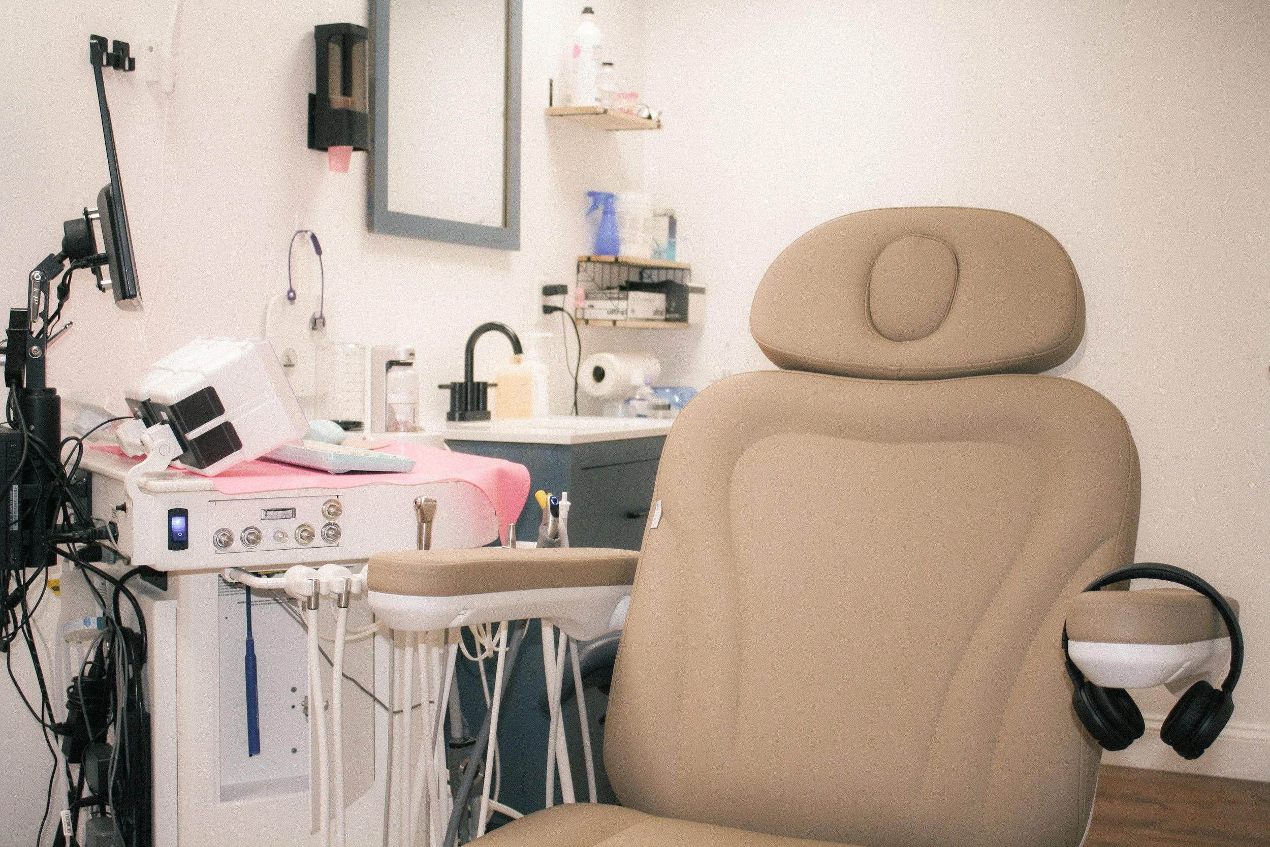 A medical examination room features a beige examination chair and a side stool. Medical equipment is present, including a monitor, a white medical device on a mobile stand, and various bottles and supplies on a shelf and countertop. The room has a neutral wall color, with some electrical cords and outlets visible.