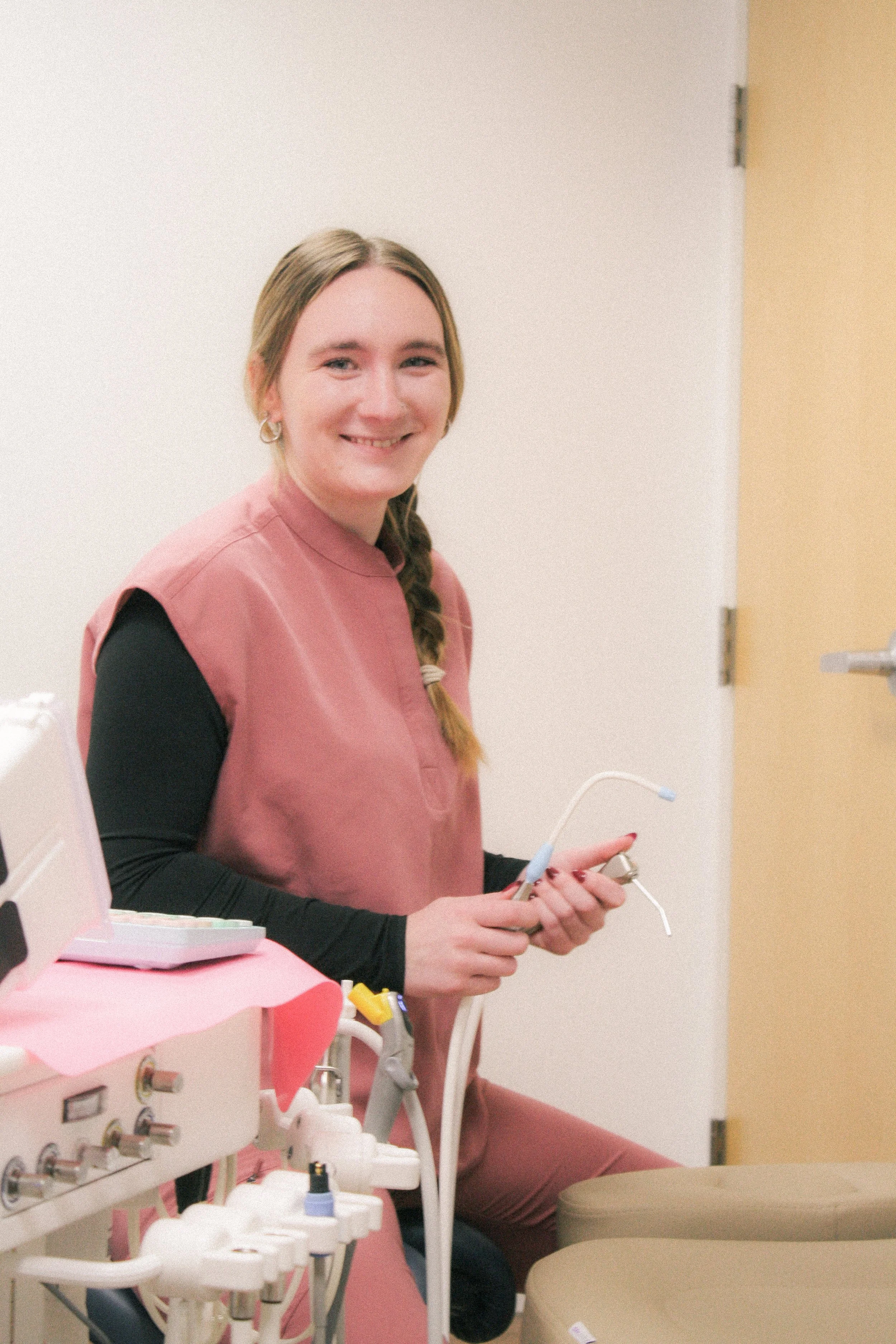 Austin with long braided hair wearing a pink sleeveless top and earrings, holding dental tools, sitting in a dental clinic.
