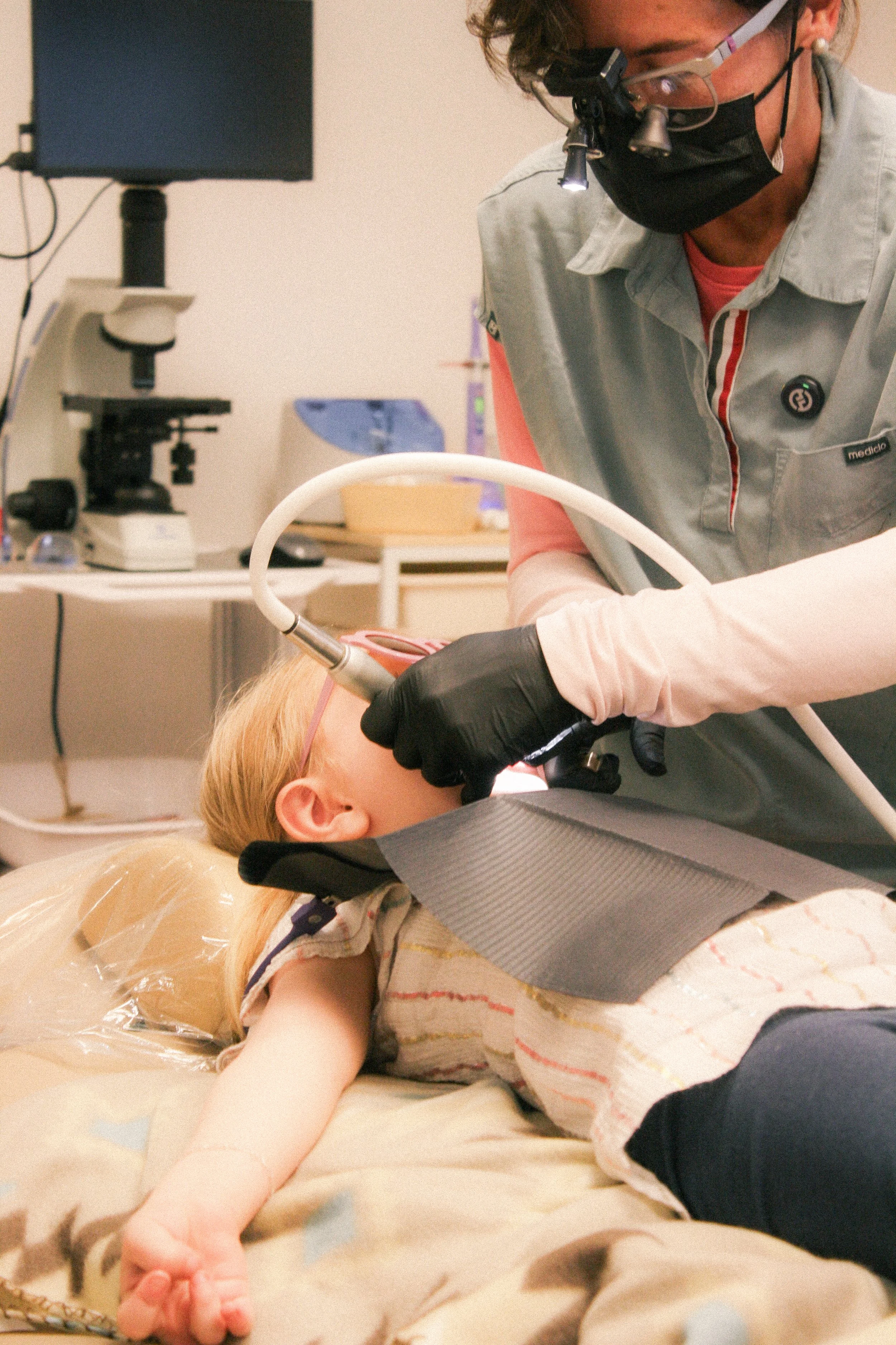 Medical professional wearing magnifying glasses and a face mask performing a procedure on a young girl lying on a medical bed, with medical equipment in the background.