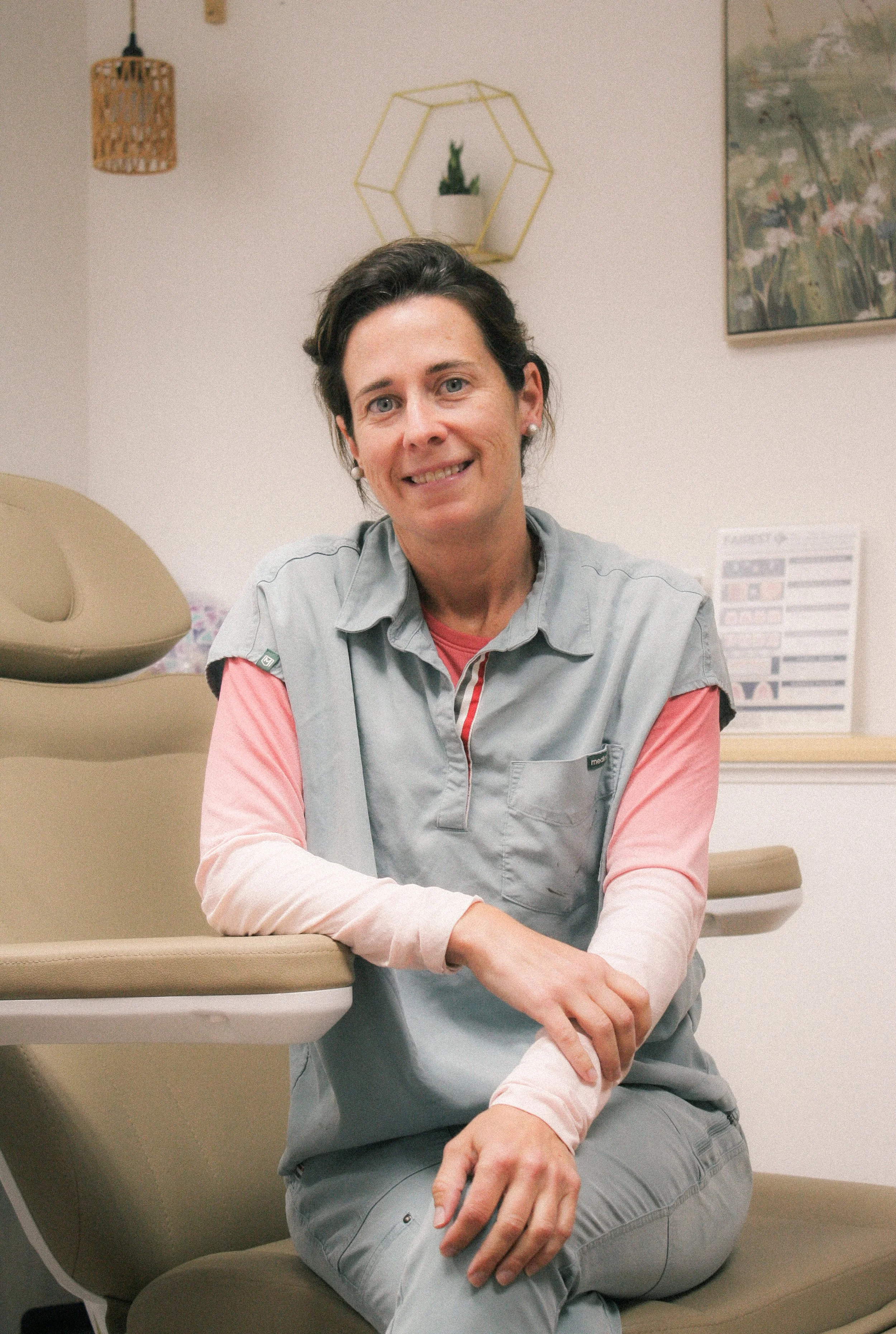 Owner and Founder, Kala French, wearing a gray medical uniform with pink sleeves, sitting in a medical examination room, smiling at the camera.