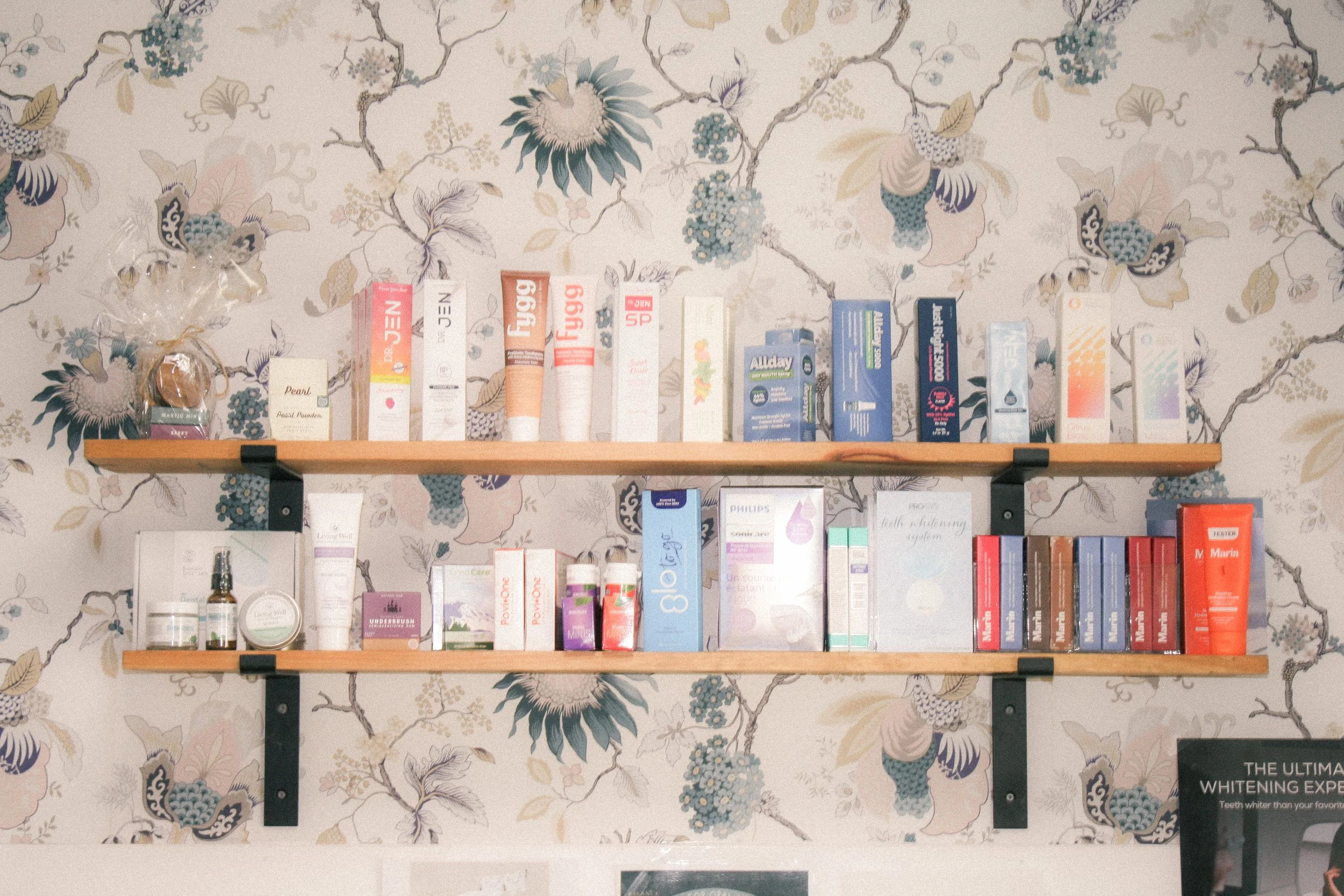 A wooden shelf mounted on a floral wallpapered wall displays various skincare, beauty, and health products.