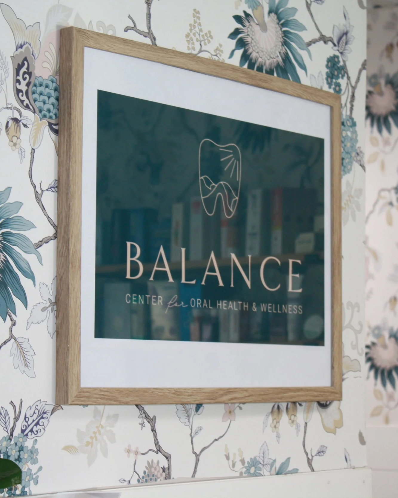 A framed poster on a wall with floral wallpaper background. The poster displays the logo of 'Balance Center for Oral Health & Wellness' with a tooth icon and text.