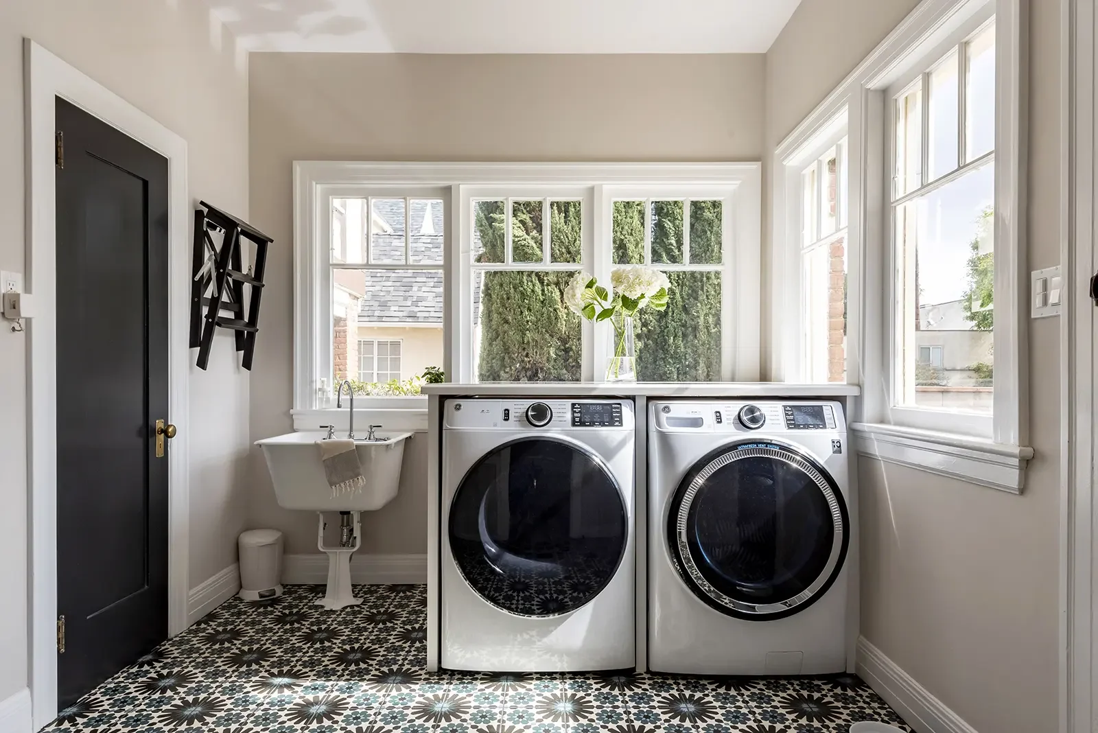 Laundry room with washer and dryer, a small sink, a window with flower vase, patterned tile floor, dark door, and black wall-mounted coat rack.