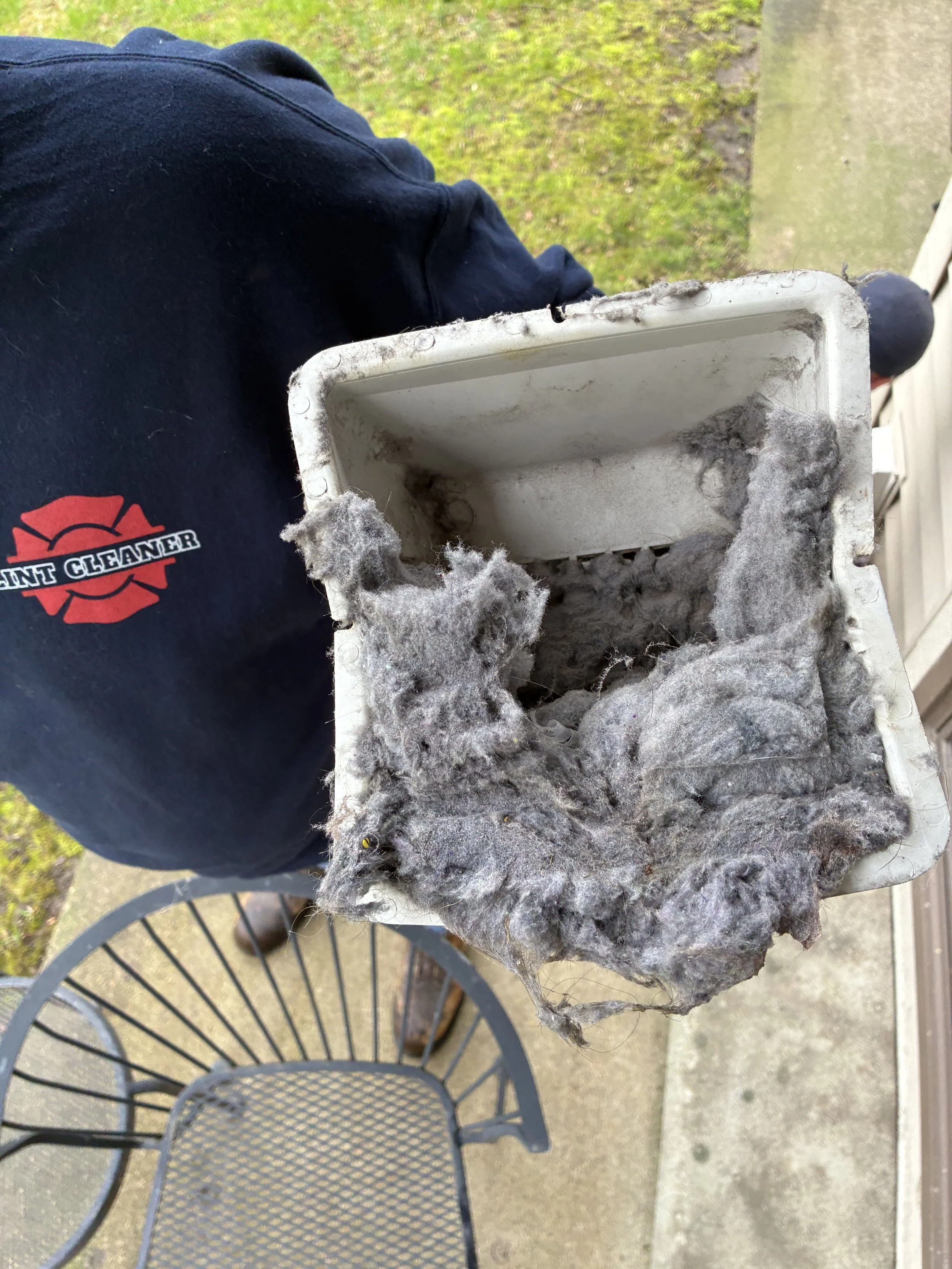 A person wearing a black shirt with a logo that says 'Paint Cleaner' is holding a dirty air filter covered in dust and debris. The background shows grass and a patio area with a metal mesh outdoor table.