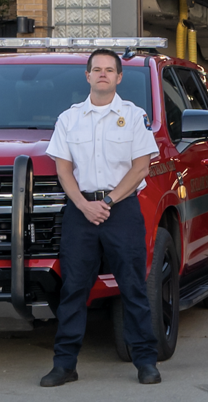 A male firefighter in uniform standing in front of a red fire truck.
