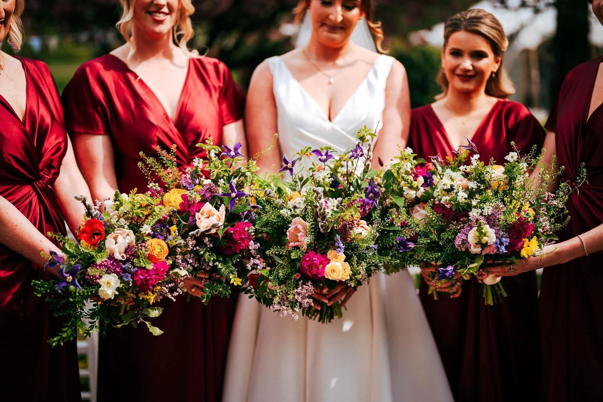 A bride dressed in white holding a large colorful bouquet of flowers, standing among bridesmaids dressed in matching red dresses, during an outdoor wedding.