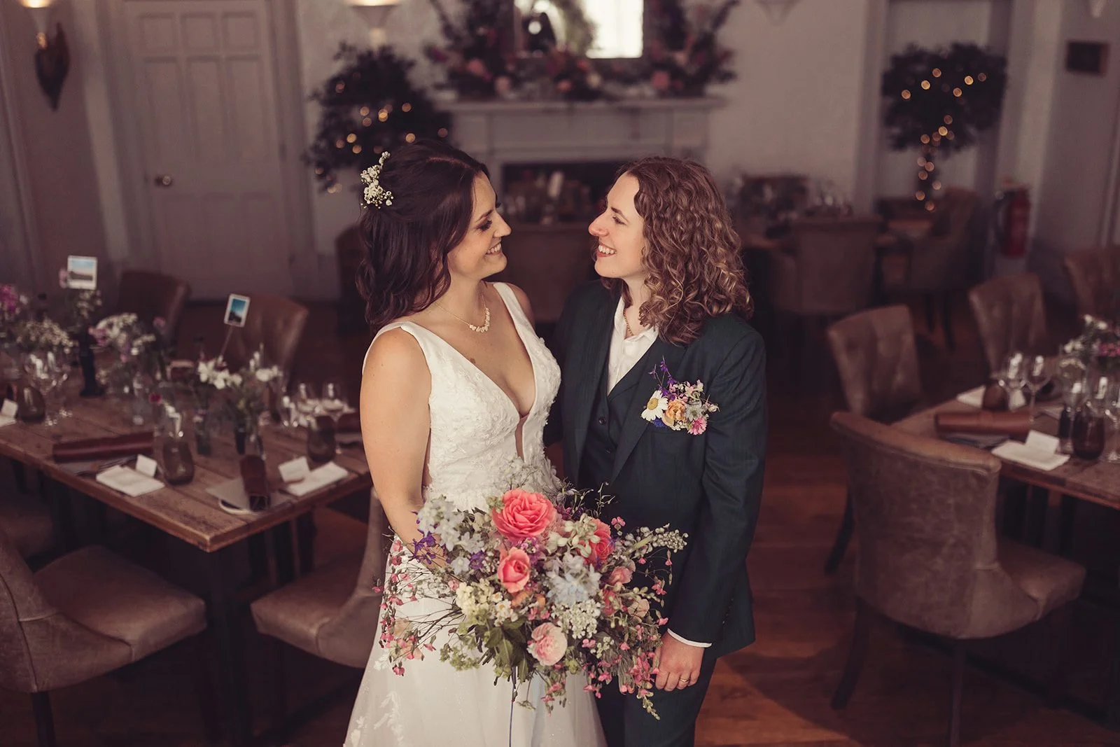 Two women, one in a wedding dress and the other in a suit, stand close together and smile at each other in a decorated event space with tables and chairs in the background.
