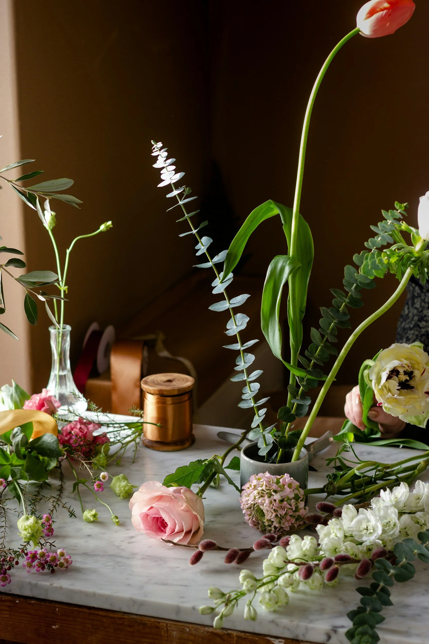 Various fresh flowers and greenery arranged on a marble surface, with some scattered around, and a person partially visible on the right side holding a bouquet.