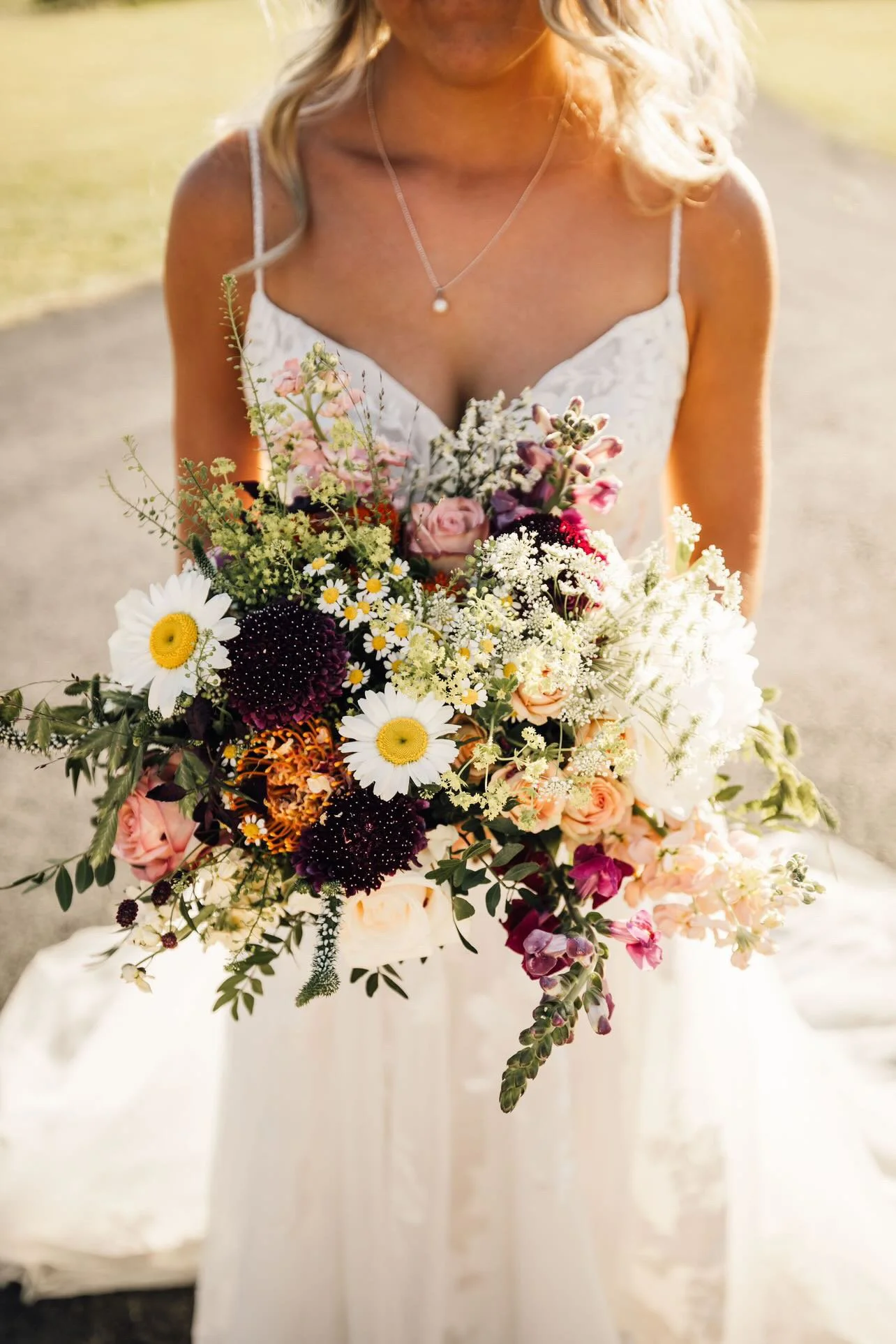 A woman in a white dress holding a large bouquet of mixed flowers, including daisies, roses, and other wildflowers, outdoors on a sunny day.