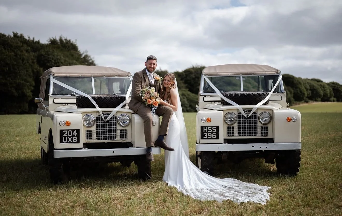 A bride and groom sitting on the front of a vintage off-road vehicle, smiling, with trees and an open grassy field in the background, during their wedding.
