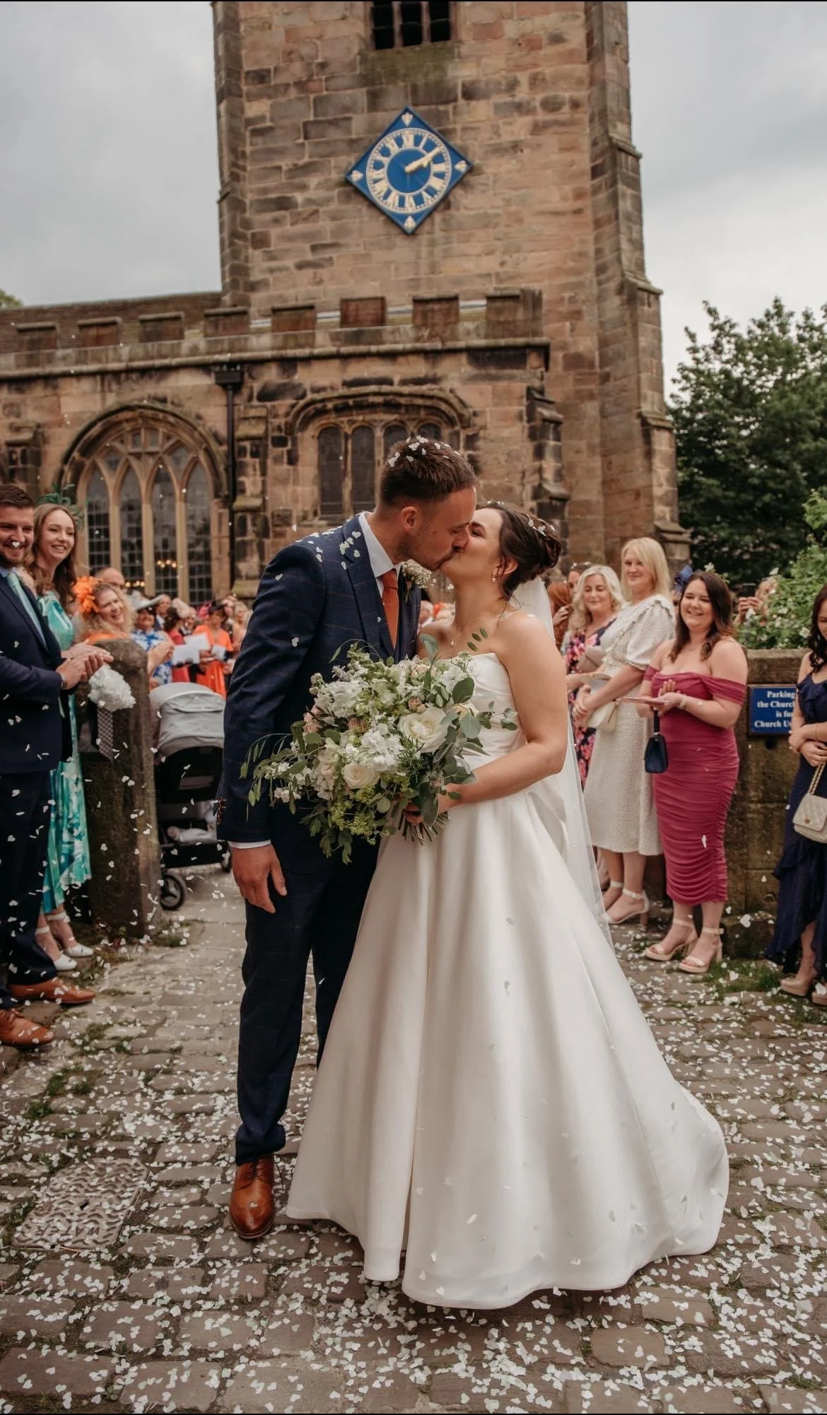 A newlywed couple, a man in a dark suit and a woman in a white wedding dress, kiss during their outdoor wedding ceremony in front of a stone church. The bride holds a large bouquet of white and green flowers. Guests in colorful attire stand around, s