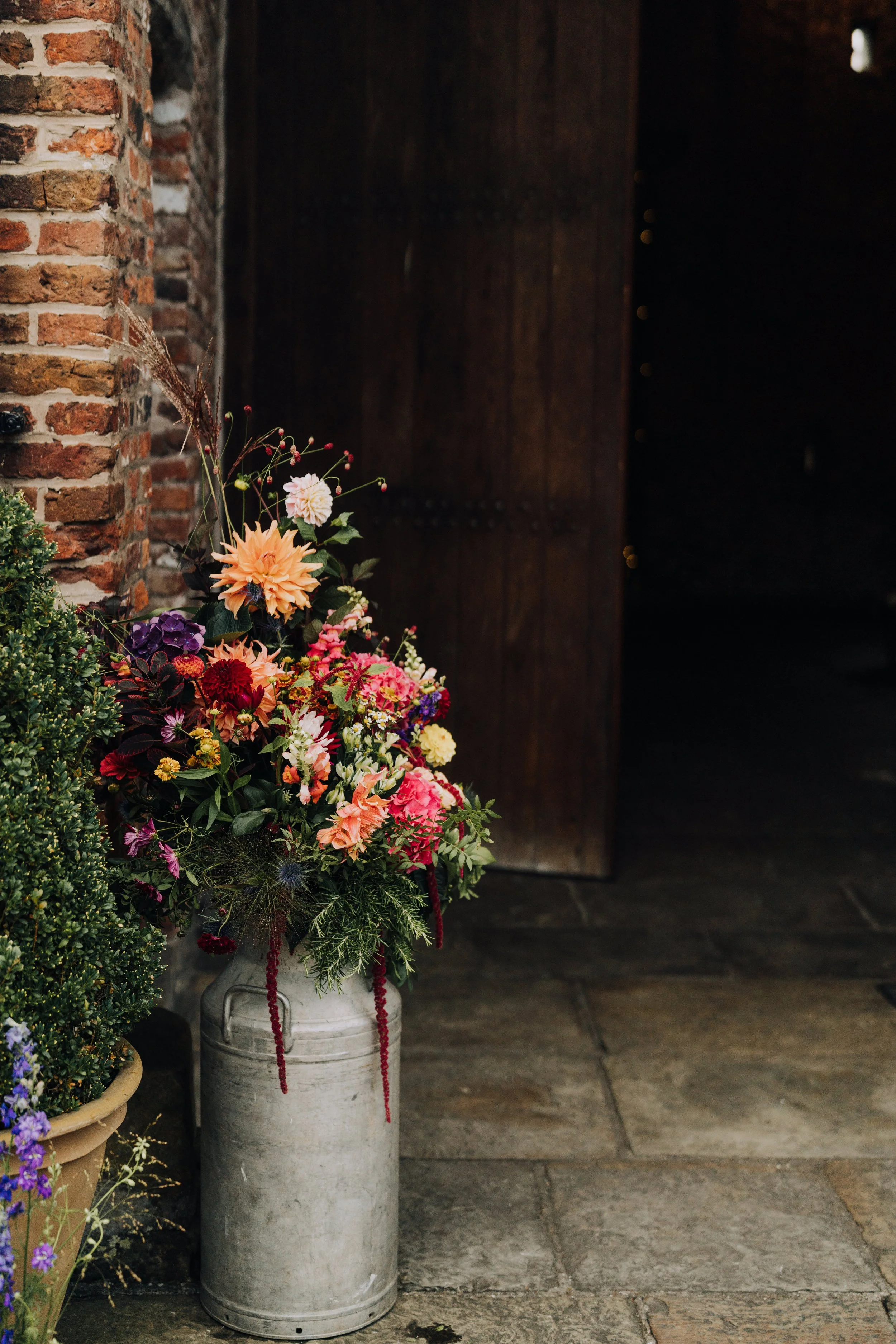 A bouquet of mixed colorful flowers in a metal bucket placed on a stone floor near a brick wall and a dark wooden door.