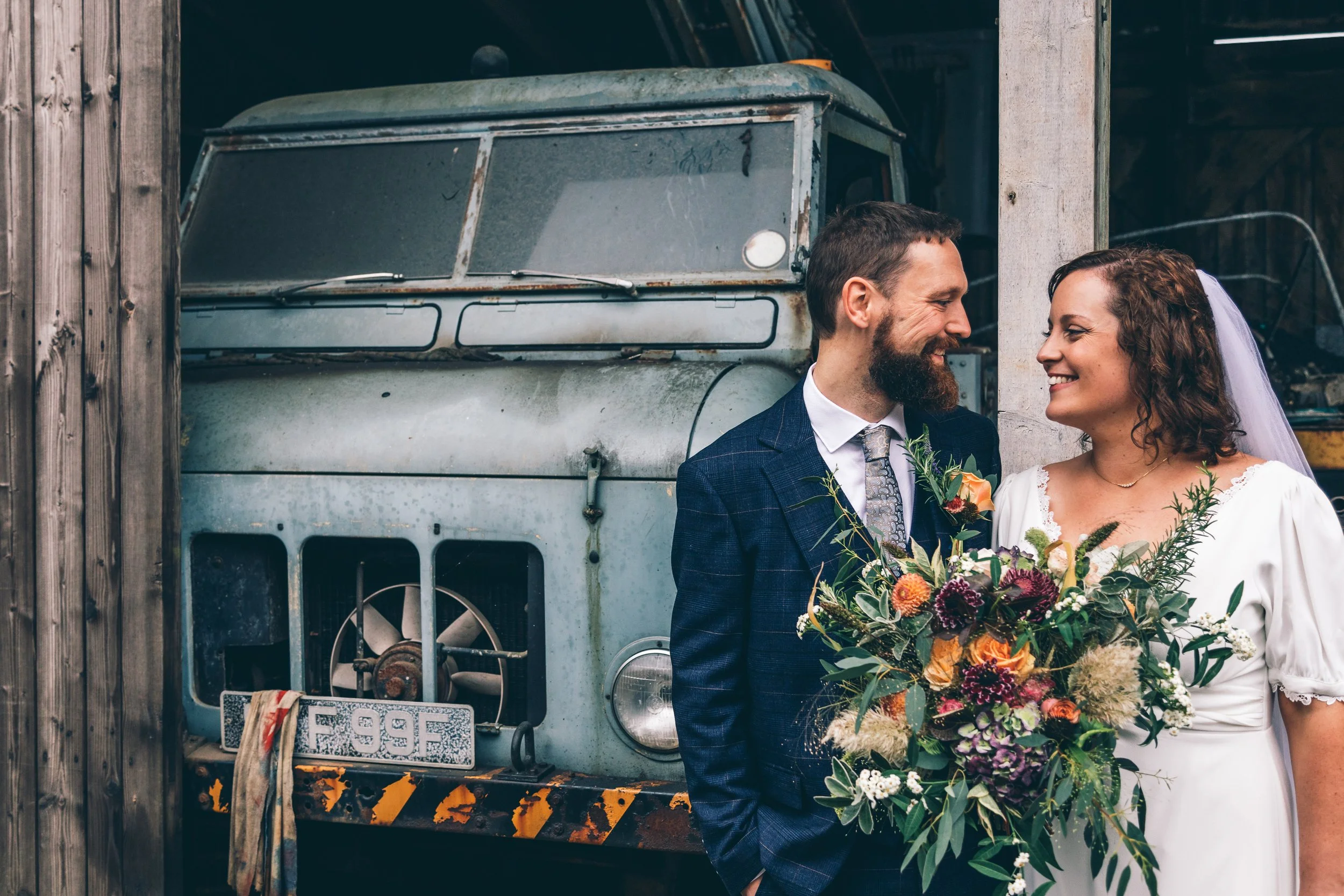 A bride and groom smiling at each other, standing in front of a rustic old vehicle, holding a large bouquet of colorful flowers.