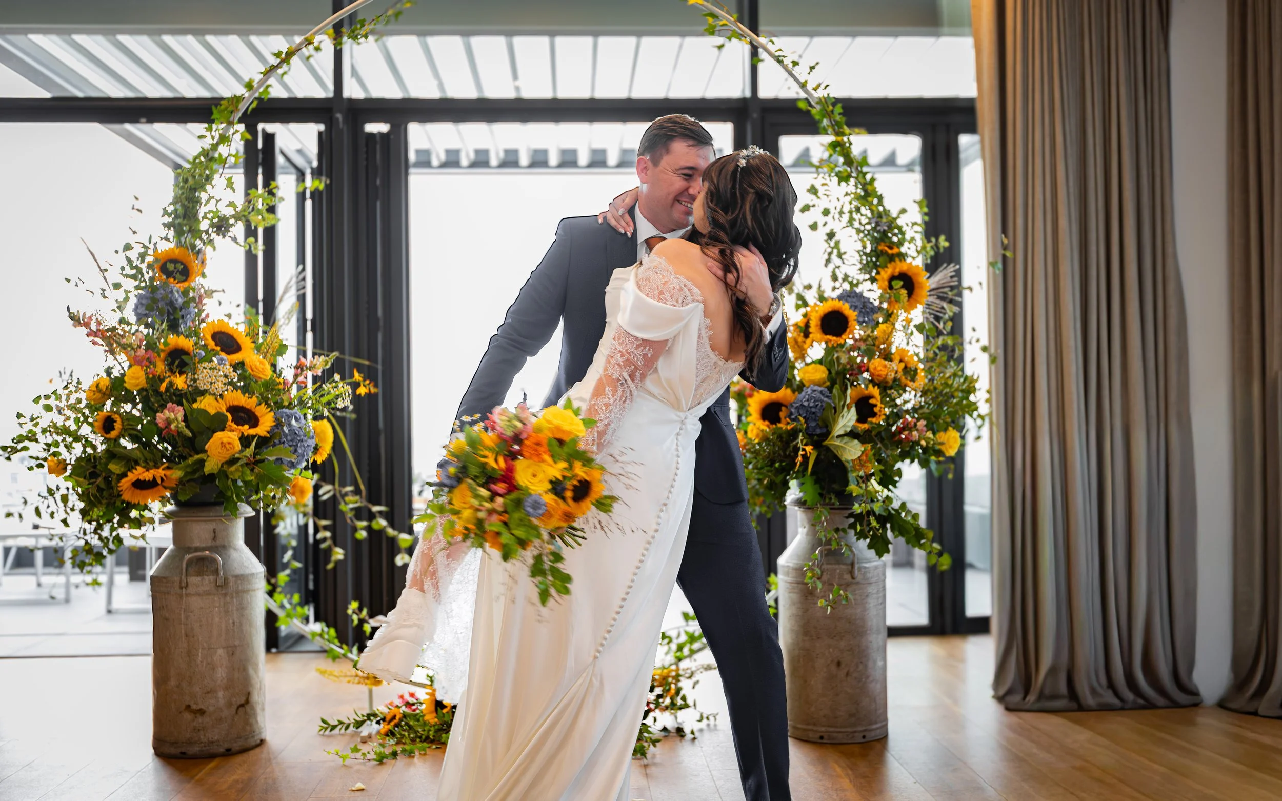 A bride and groom sharing a kiss during their wedding ceremony, surrounded by sunflowers and other colorful flowers in vases, inside a modern venue with large windows and gold curtains.