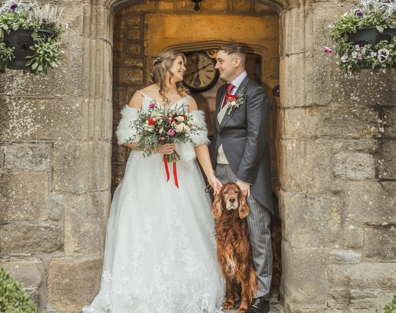 A bride and groom standing in a stone archway, smiling at each other, with a brown dog between them. The bride holds a bouquet and wears a white wedding dress with fur shawl, while the groom is in a gray suit with a red tie and boutonniere. There are